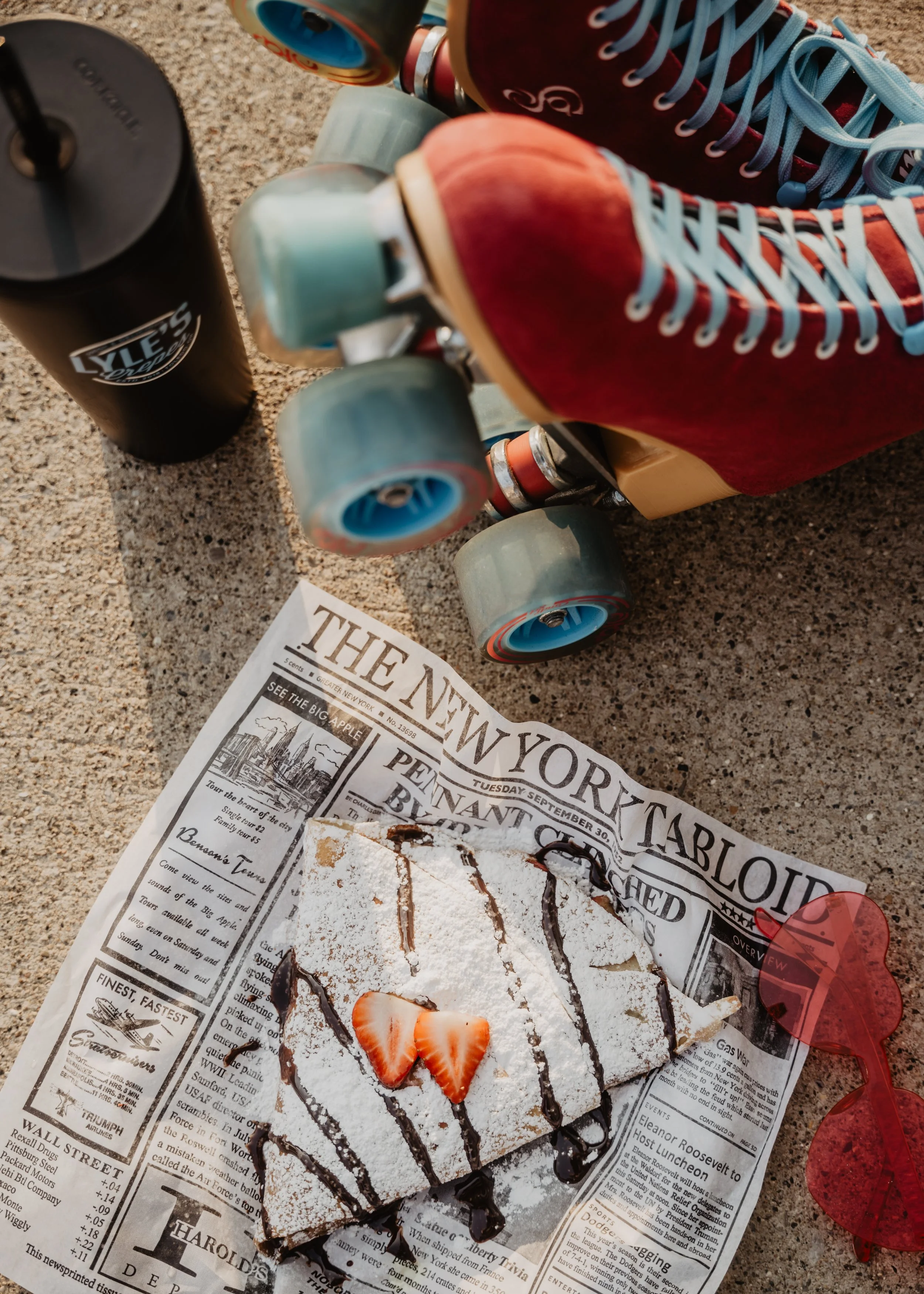 A slice of chocolate and strawberry cake topped with sliced strawberries, drizzled with chocolate syrup, on a newspaper background. Nearby, a pair of red sunglasses, a black water bottle, skateboards with colorful wheels, a paper with printed text, a