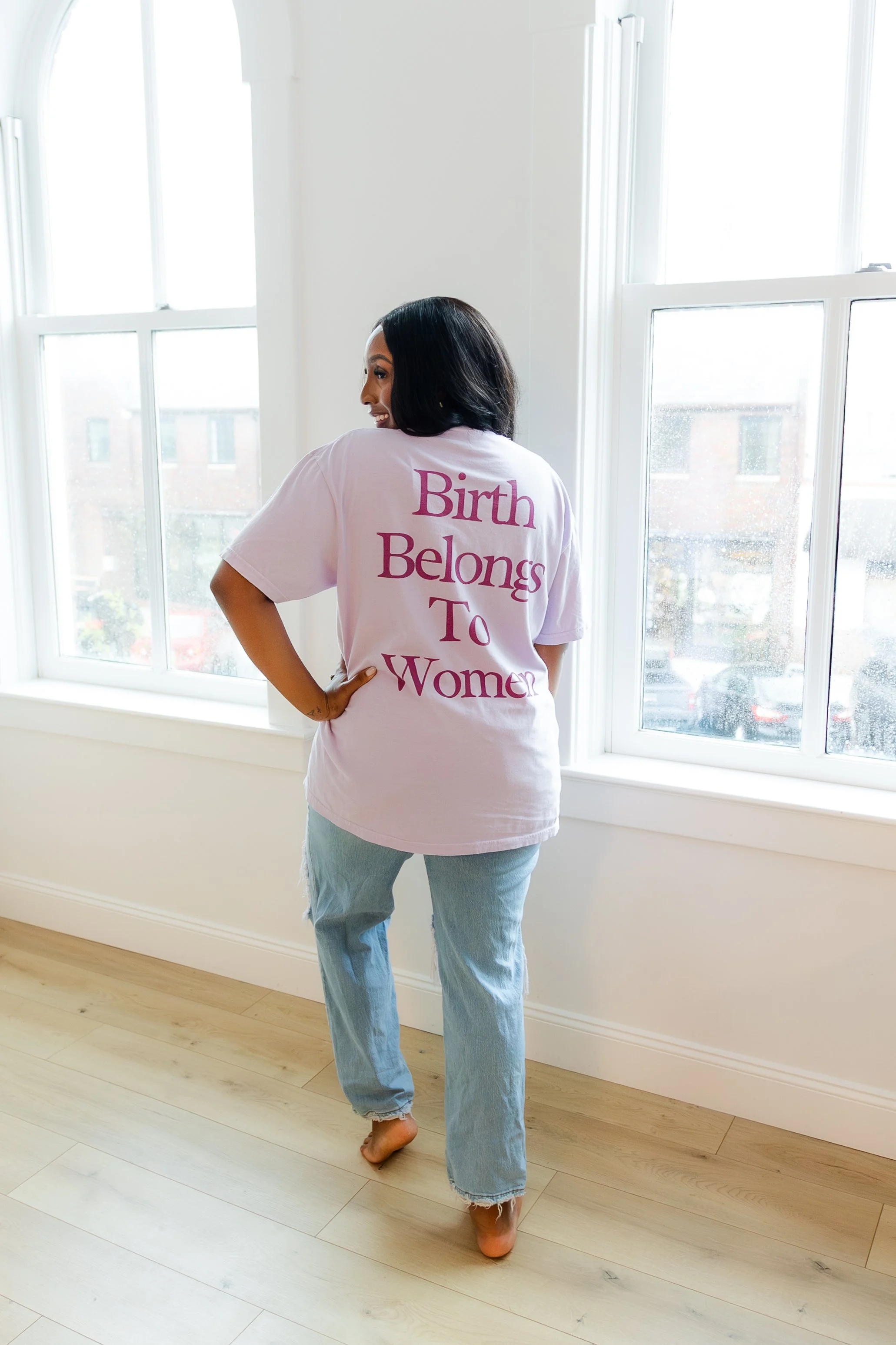 Woman standing indoors near large windows, wearing a light pink t-shirt with the text "Birth Belongs To Women" on the back, and light-wash jeans.