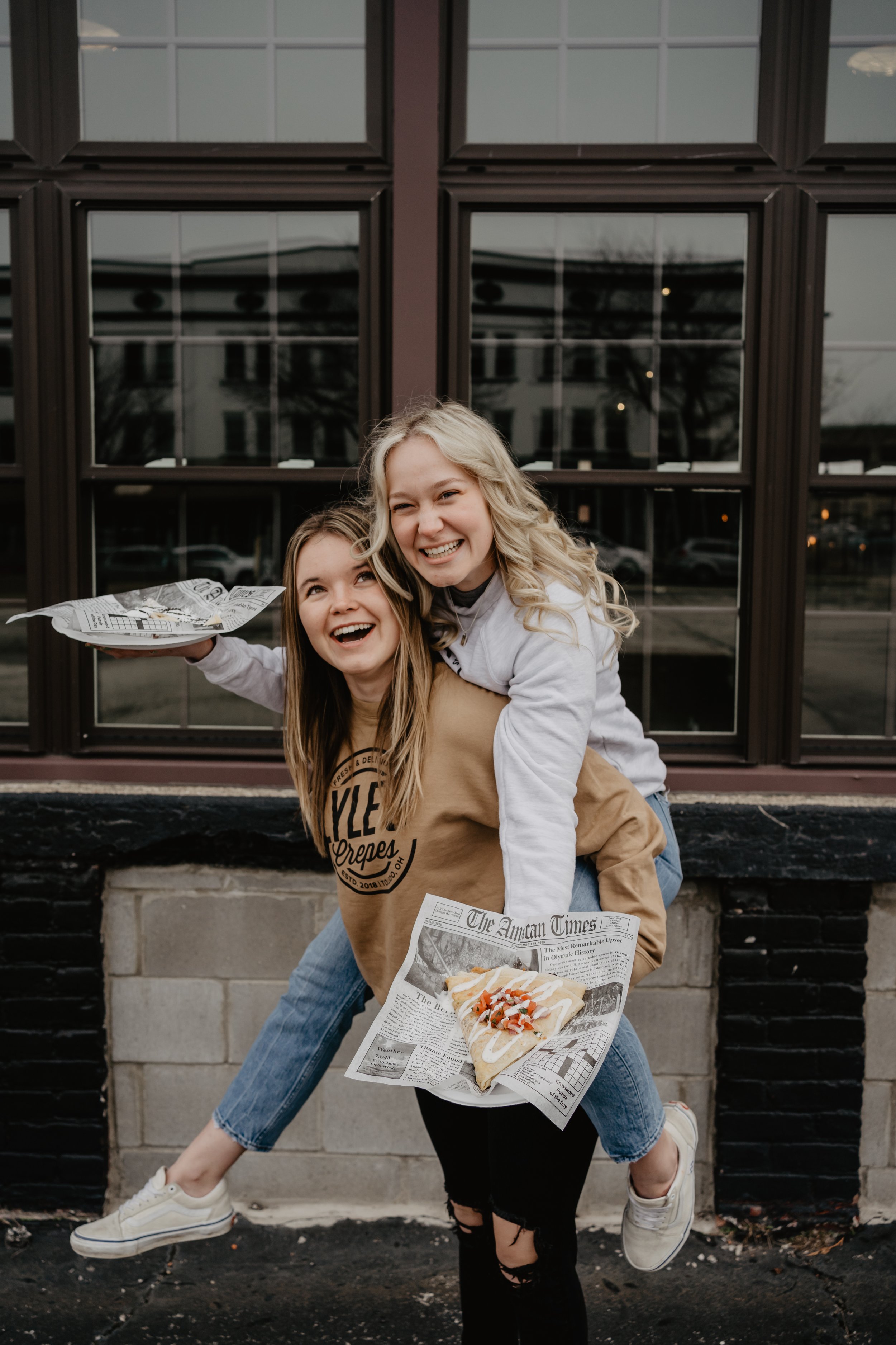 Two young women smiling and playing outside in front of a building with large windows. One woman is giving the other a piggyback ride while holding a newspaper and a slice of pizza. The scene appears fun and playful.