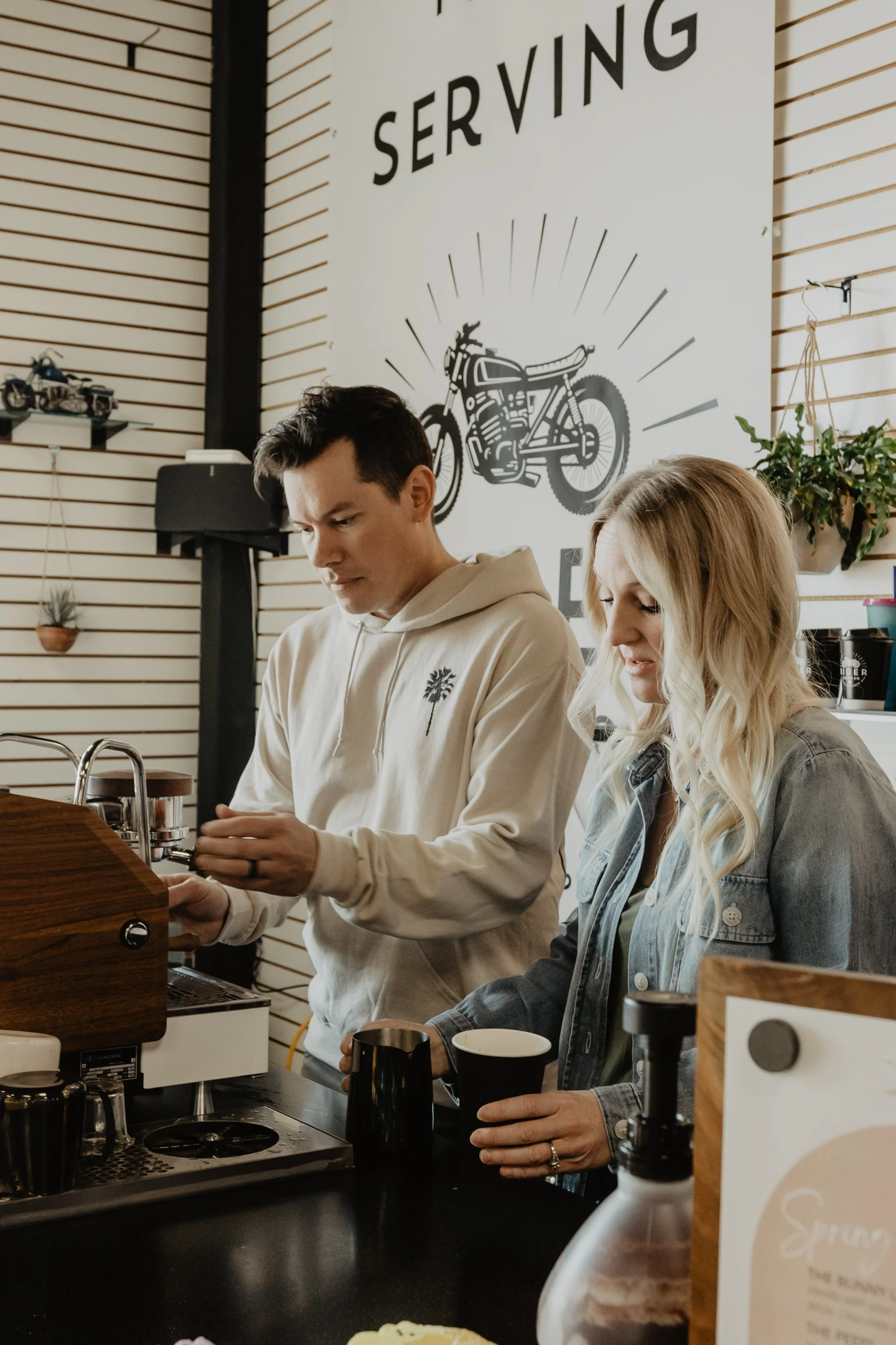 Two people preparing coffee behind a counter in a coffee shop, with a sign featuring a motorcycle and the word 'SERVING' in the background.