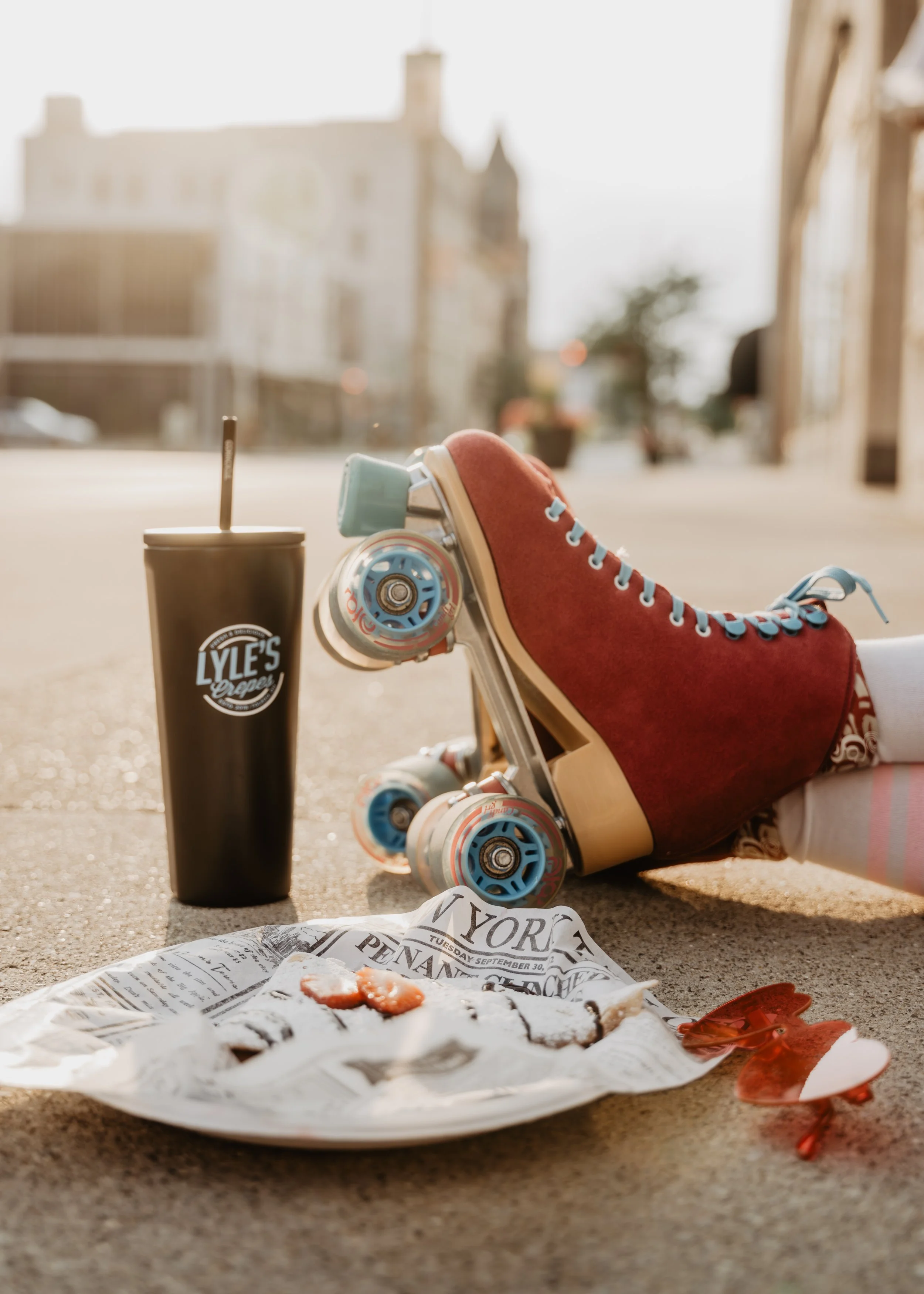 A skateboarding shoe, a soda cup with a straw, a crumpled newspaper, and some food in a wrapper on the ground, with a blurred urban background