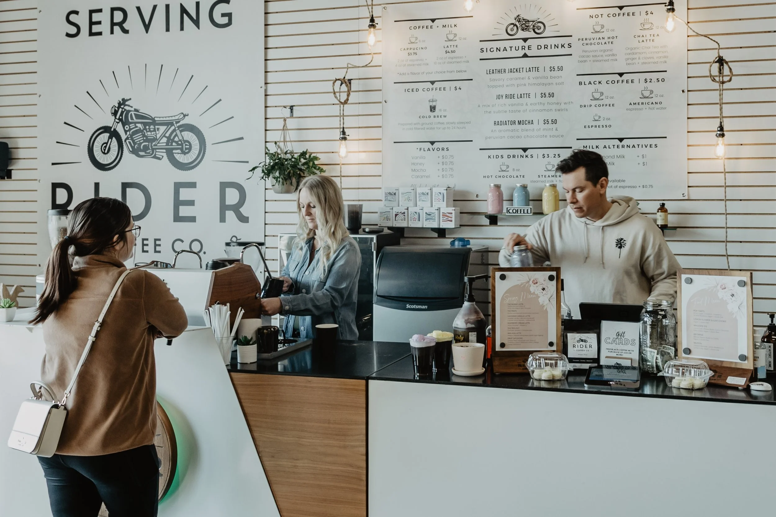 Customers ordering at a modern coffee shop counter with a large sign. The staff is preparing drinks, and a barista is pouring a beverage. The menu displays various coffee options and prices, with rustic hanging light bulbs and a plant in the backgrou