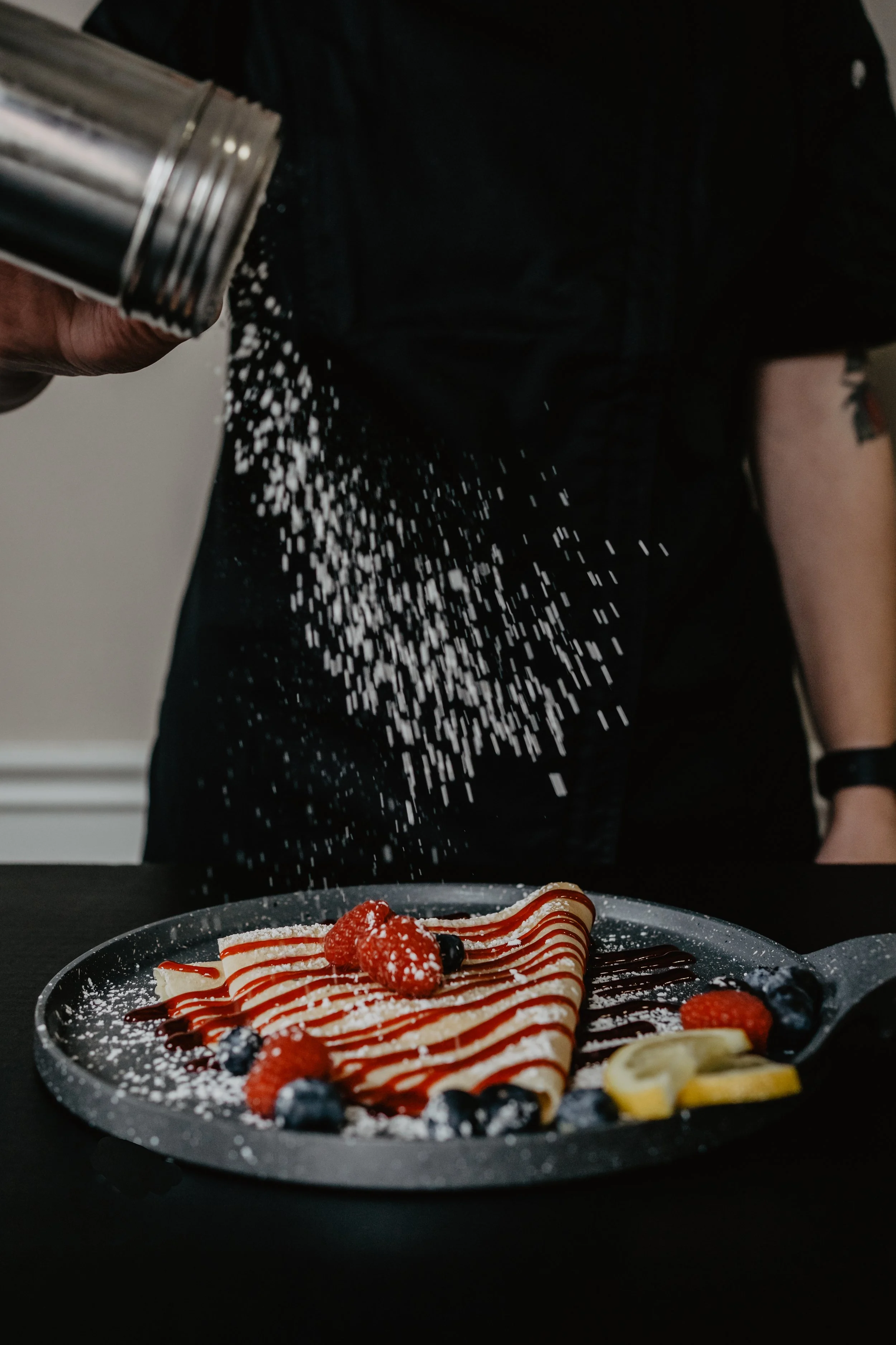 A person sprinkling powdered sugar on a dessert plate with strawberries, blueberries, and banana slices. In the background, a plate of dessert is visible, topped with a strawberry, raspberries, blueberries, and drizzled with chocolate and caramel.
