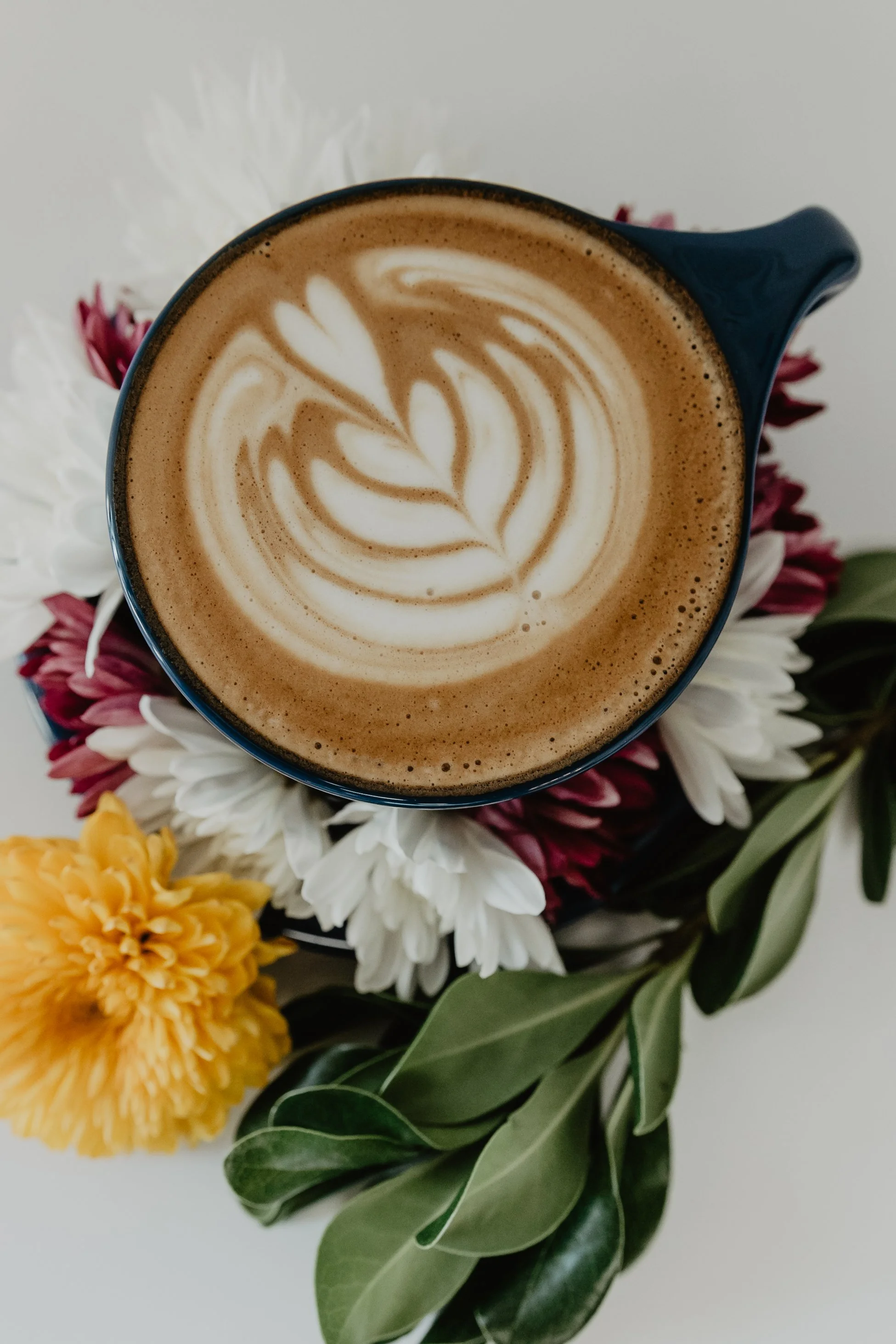 A cup of coffee with latte art resting on a bed of white, yellow, and red flowers and green leaves.