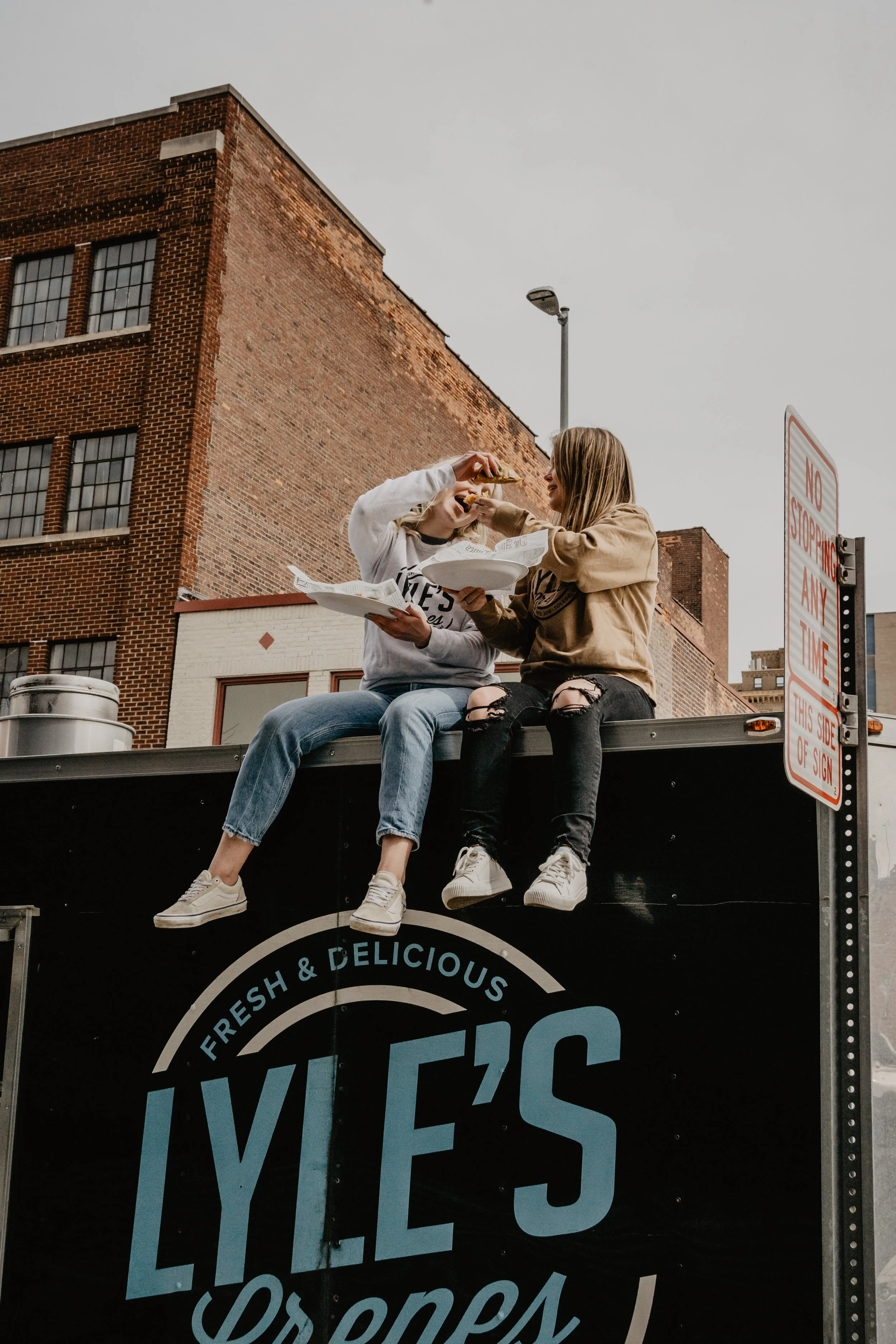 Two young women sitting on top of a food truck, sharing a slice of pizza outdoors in an urban area with brick buildings and street signs nearby.