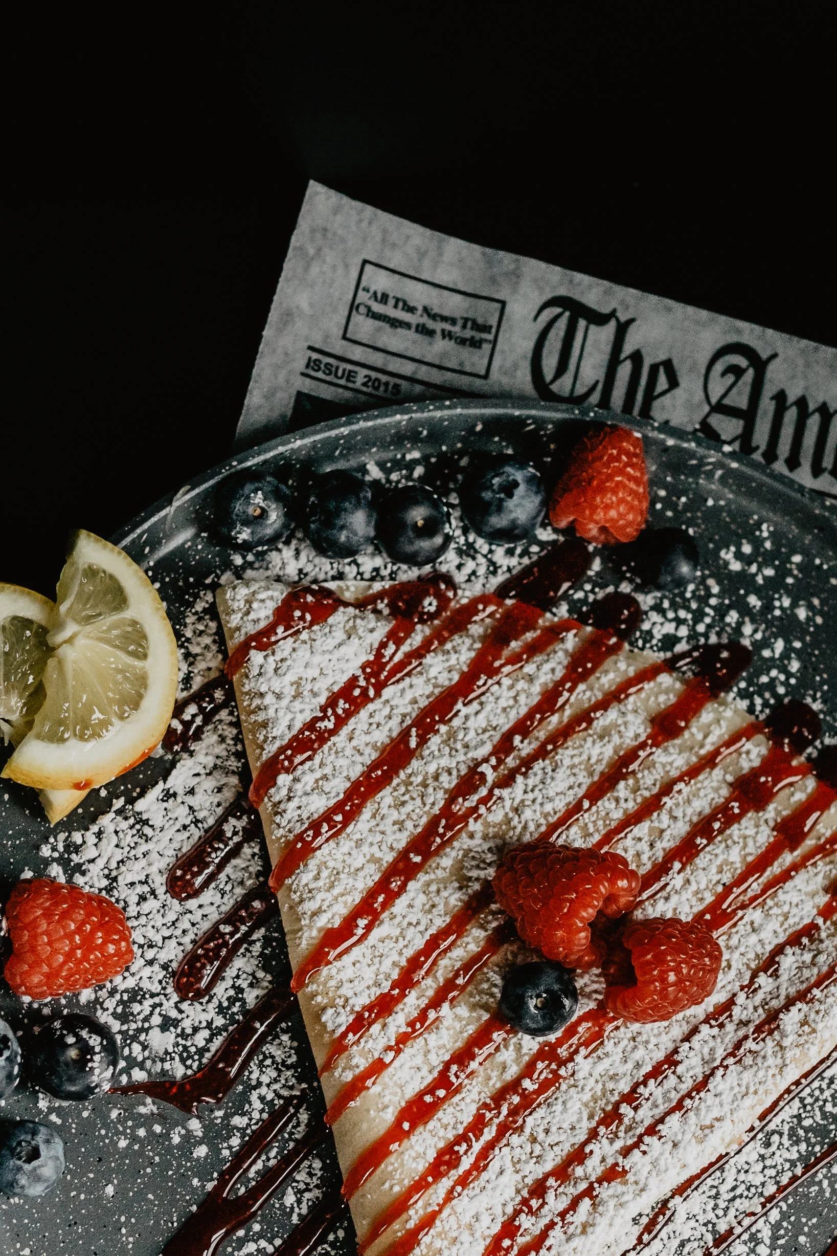 A partially visible newspaper in the background, with a slice of cake topped with berries, lemon slices, and drizzled with sauce in the foreground.