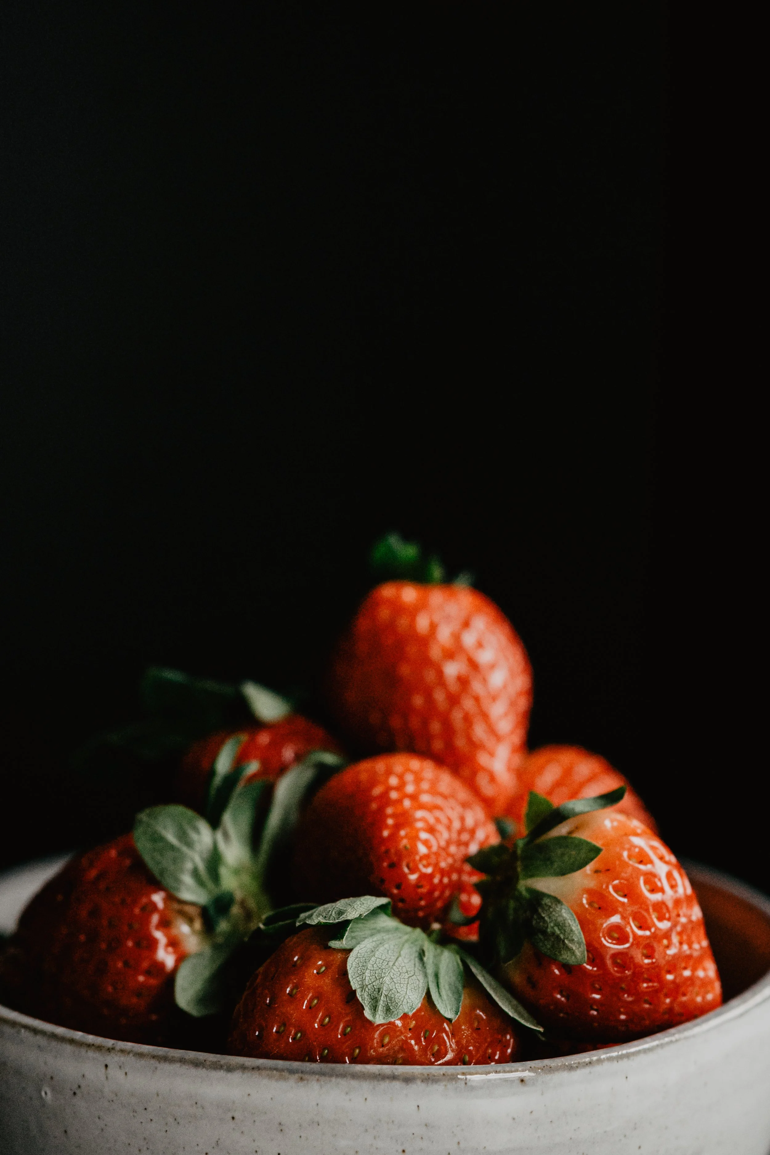 A bowl of fresh strawberries with green leaves against a dark background.