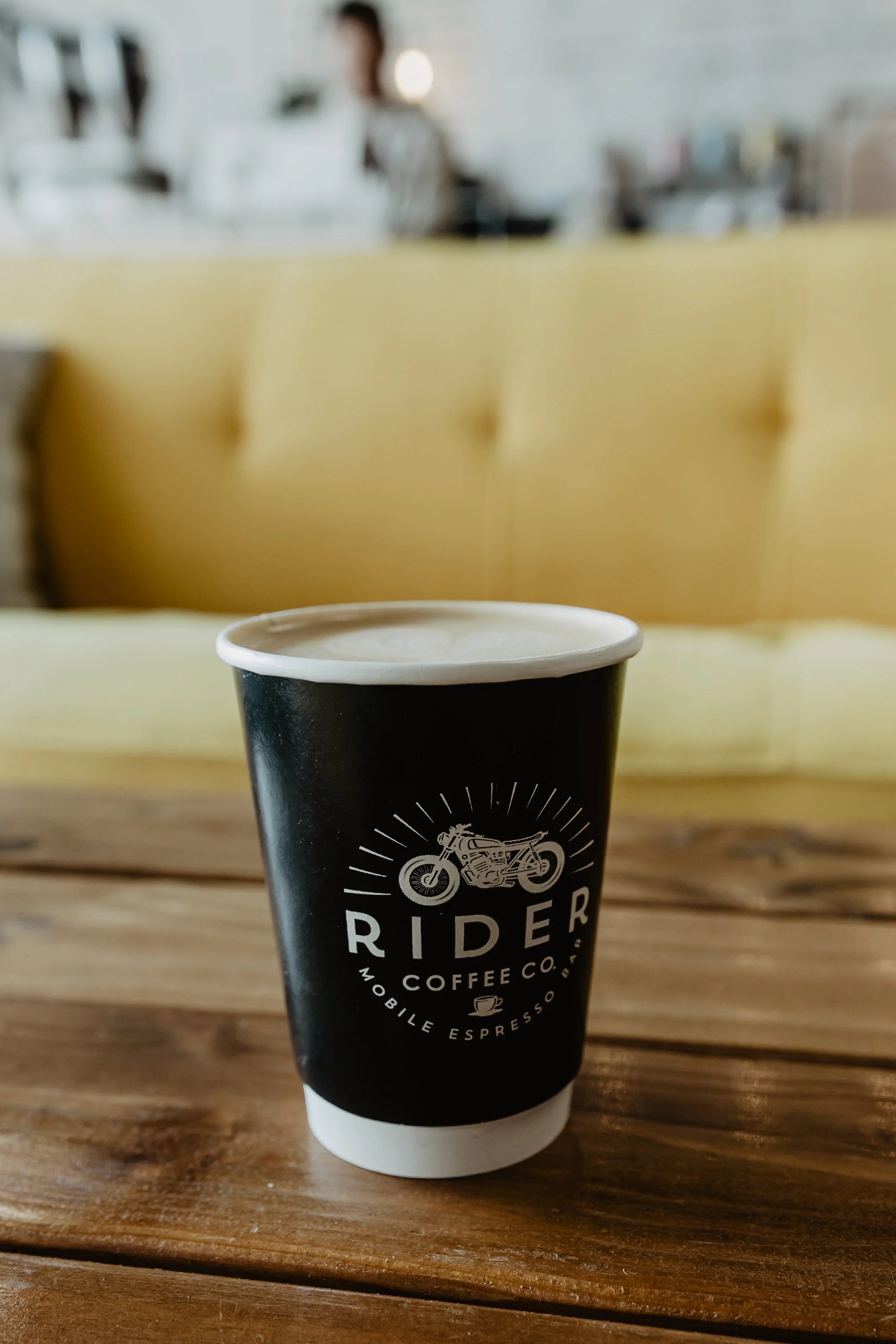 A black disposable coffee cup with a white lid sitting on a wooden table in a coffee shop. The cup has a logo of a motorcycle and the word 'RIDER' along with other text about the coffee company.