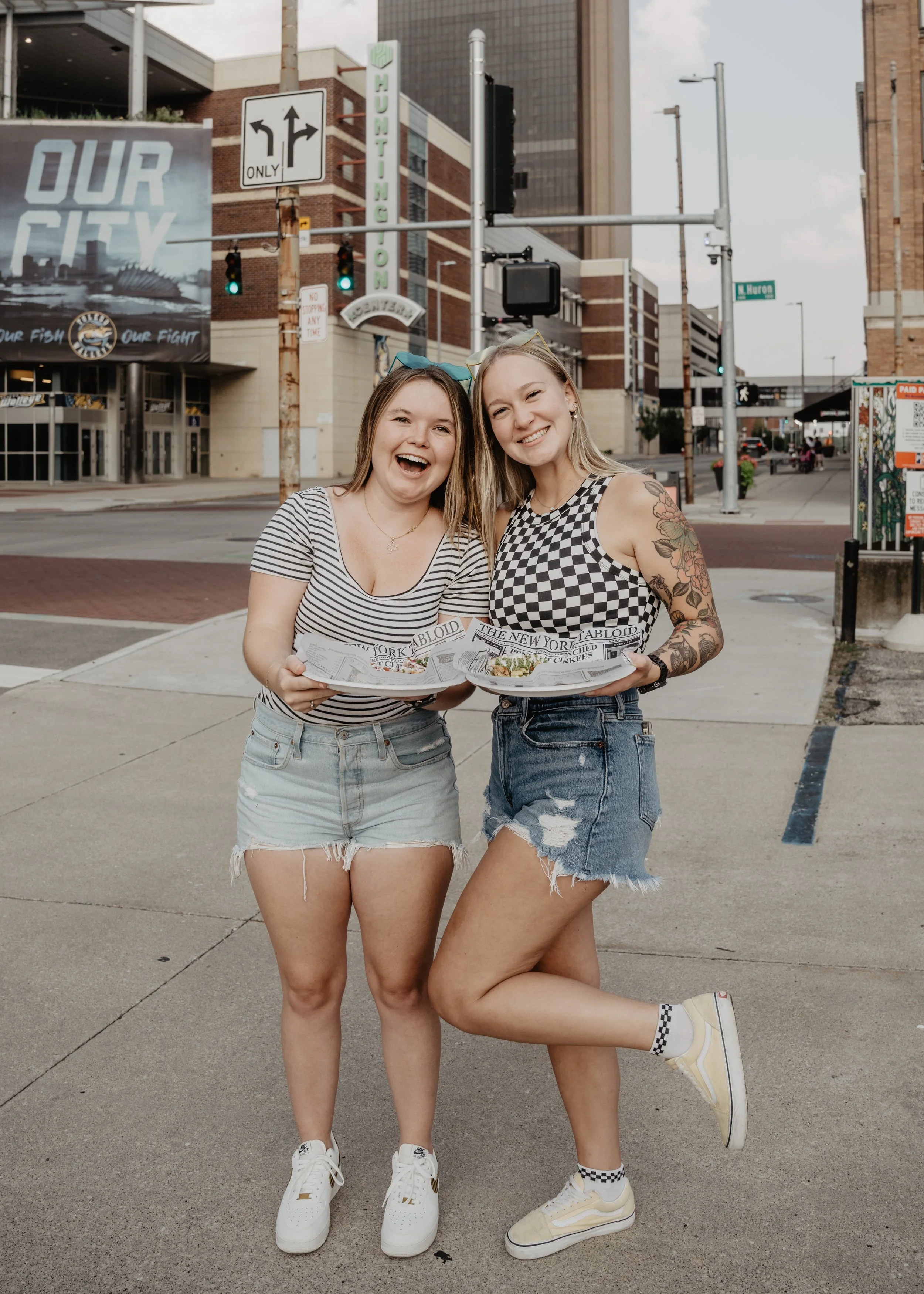 Two young women standing outside on a city street holding plates of food, smiling, one wearing a striped t-shirt and shorts, the other wearing a checkered tank top and ripped shorts, with one leg lifted in a playful pose.