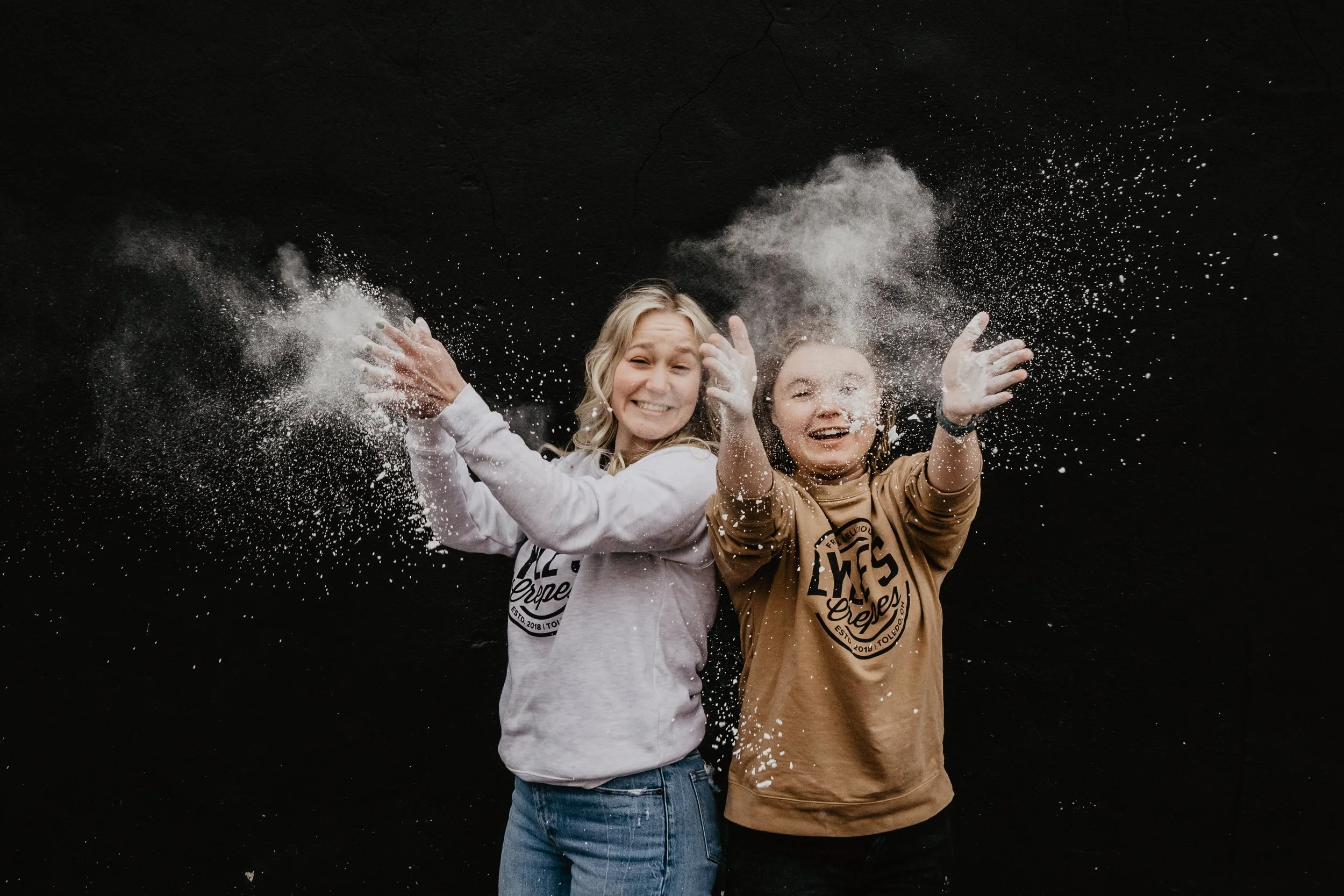 Two young women are smiling and throwing white powder or flour into the air against a black background.