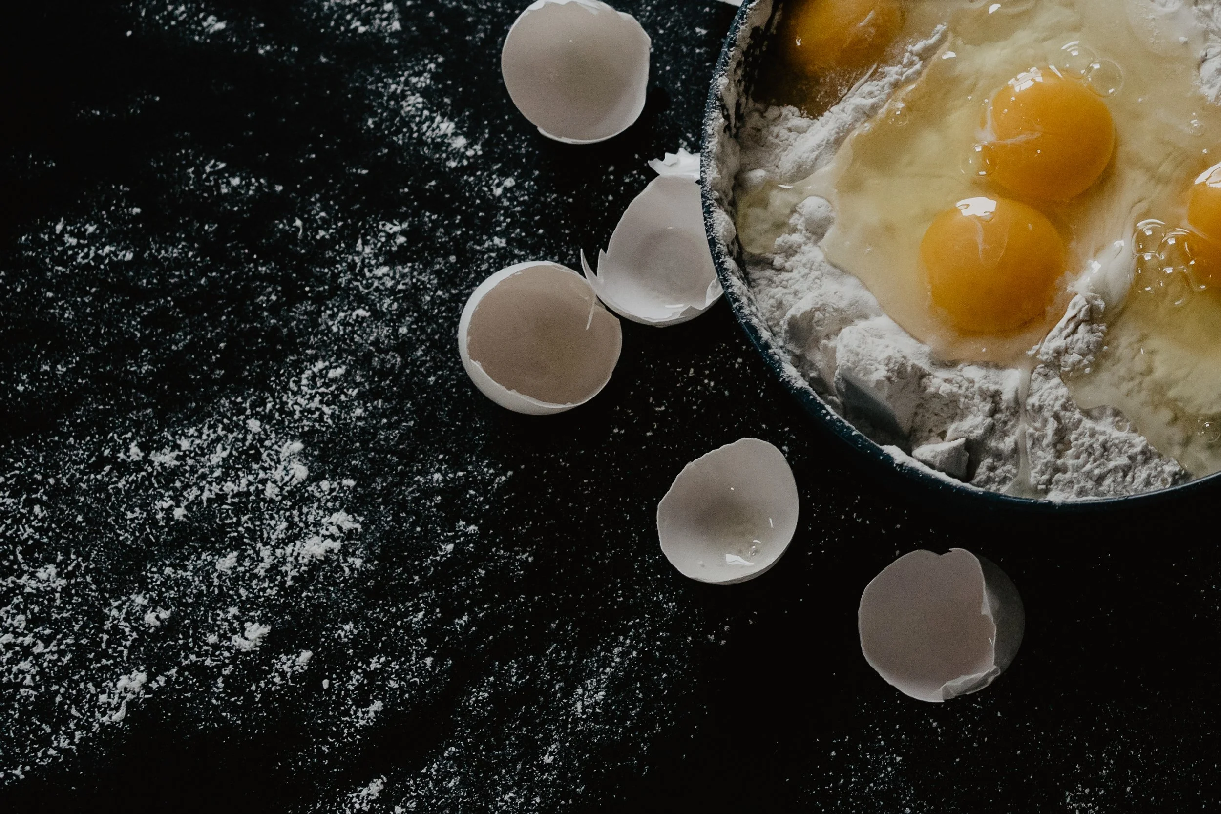 A baking mixture in a bowl with three eggs cracked into it, surrounded by broken eggshells on a black, floured surface.