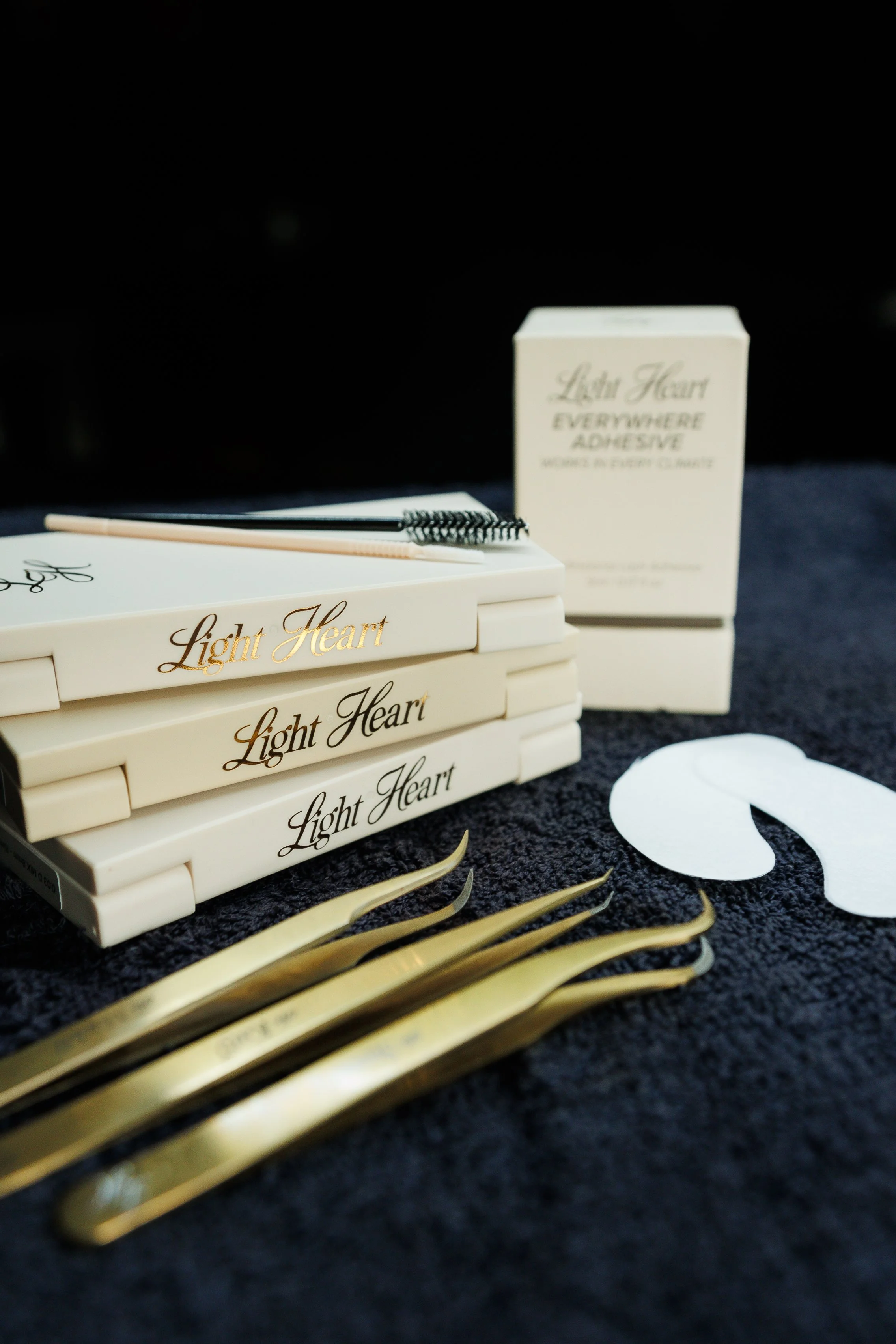 A collection of gold-colored hair dyeing tools, including brushes and combs, with a box labeled 'Light Heart Everywhere Adhesive' in the background, set on a dark surface.