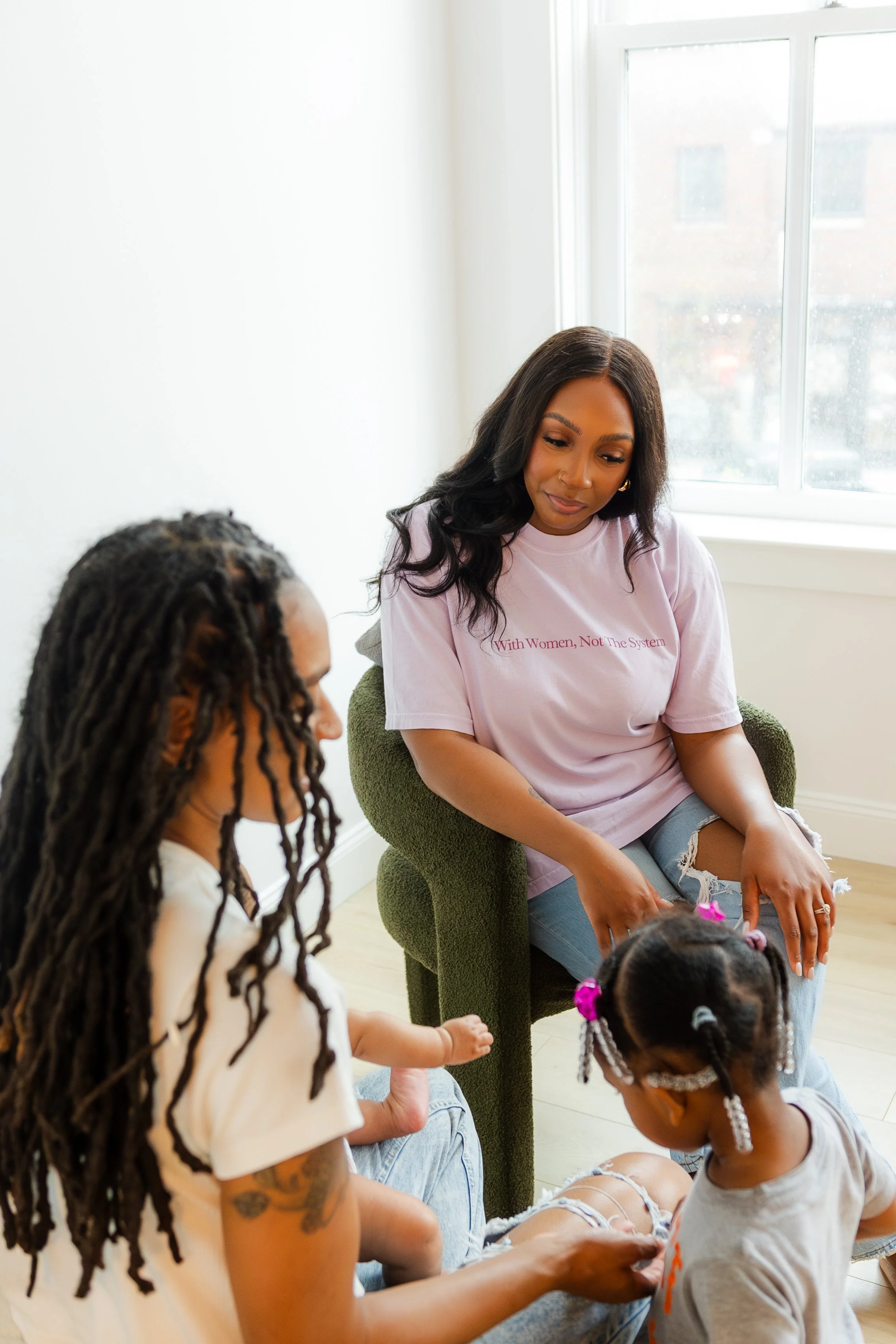 A woman with long dark hair wearing a pink T-shirt that says 'With Women, Not the System' sitting on a green chair, engaging with children in a bright room with large windows.