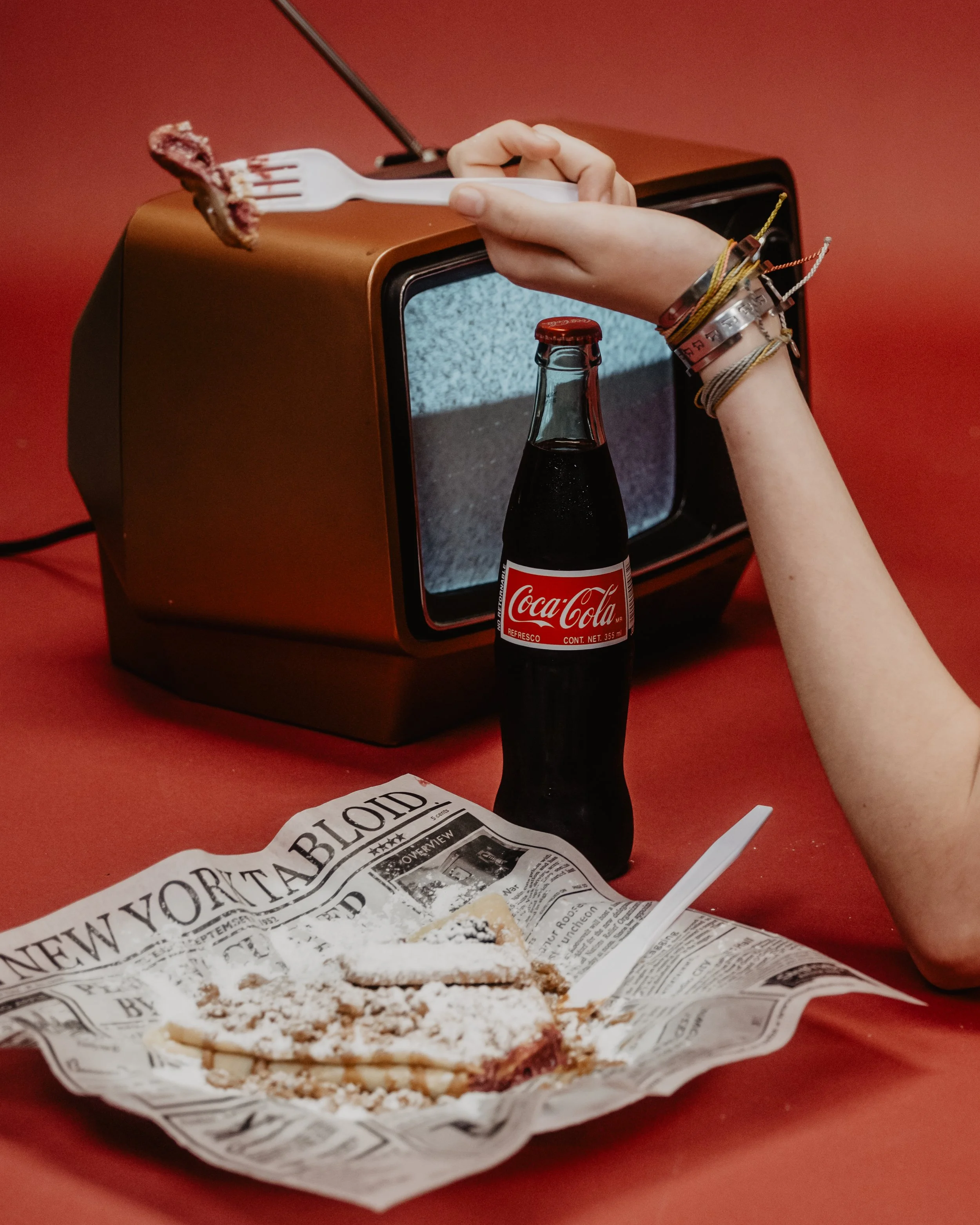 A partially eaten slice of pizza on newspaper, a bottle of Coca-Cola, a fork with cake, and a hand with bracelets in front of an old-fashioned television set.
