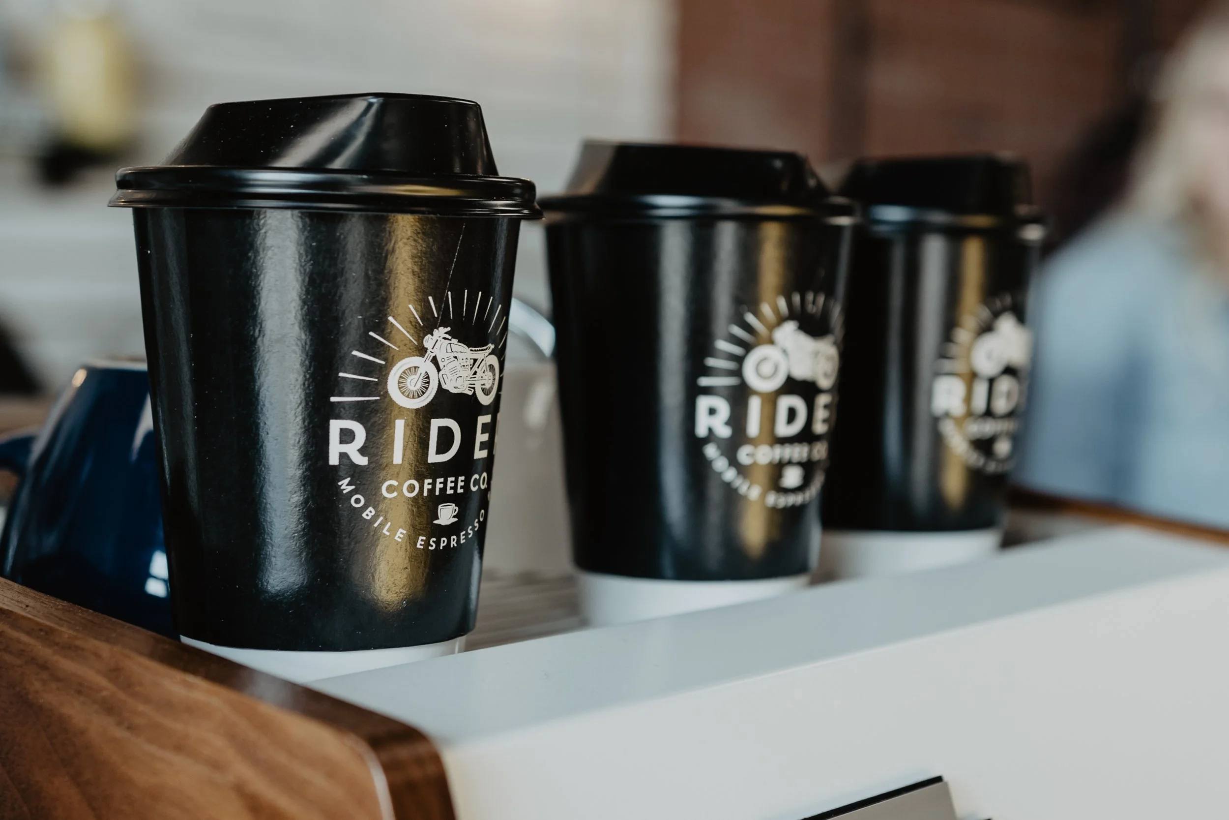 Black disposable coffee cups with a motorcycle logo and text, placed on a white shelf against a blurred background.