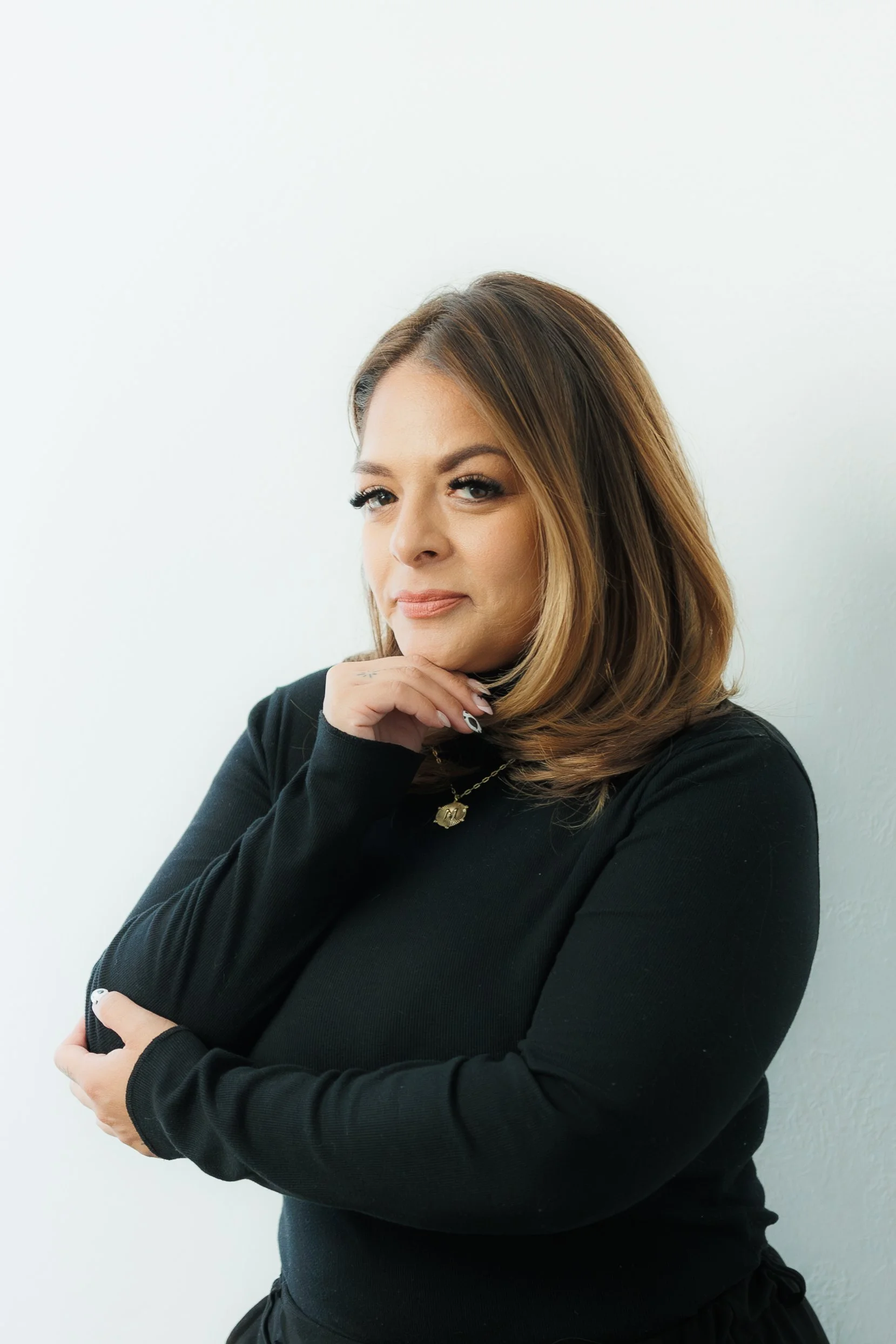 Portrait of a woman with light brown hair, wearing a black long sleeve shirt, standing against a white background, with her hand resting under her chin.