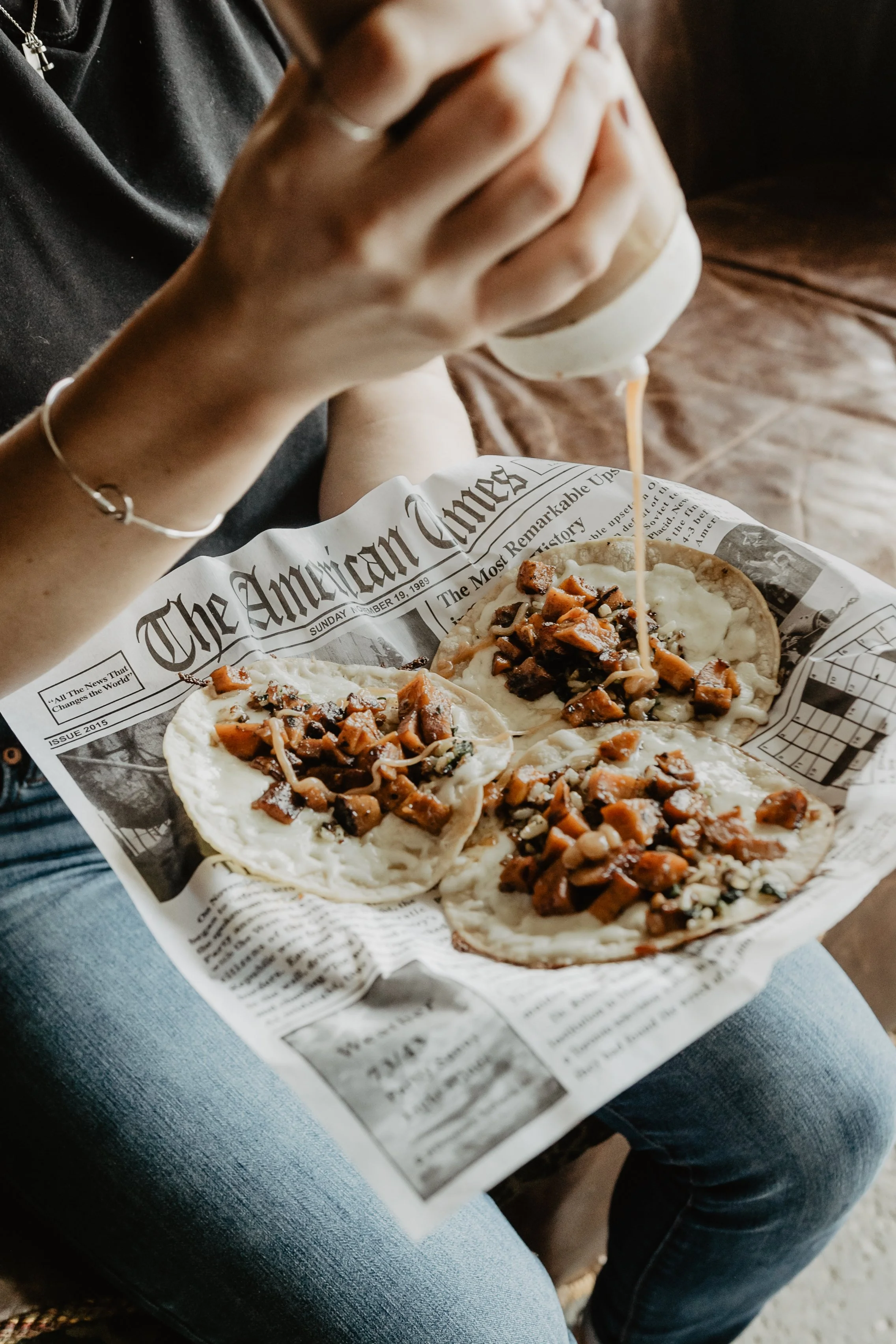 Person sitting on a blue couch, wearing blue jeans, holding a pizza with three slices and pouring hot sauce on one slice. The pizza is on a newspaper-lined tray.