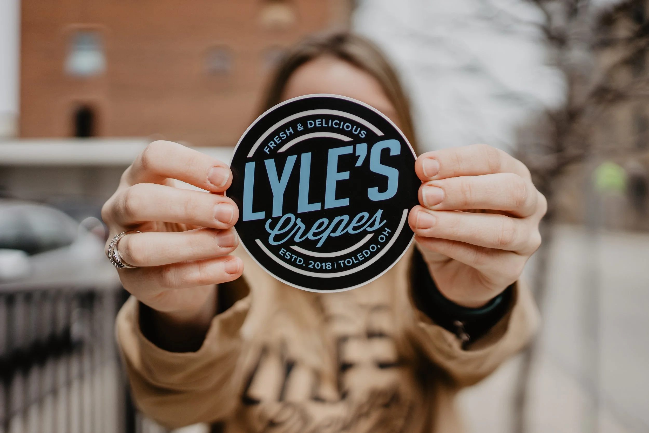Person holding a circular black and blue sticker that reads 'Lyle's Crepes' with additional text 'Fresh & Delicious' and 'Toledo, OH' in white, with a blurred background of trees and building.