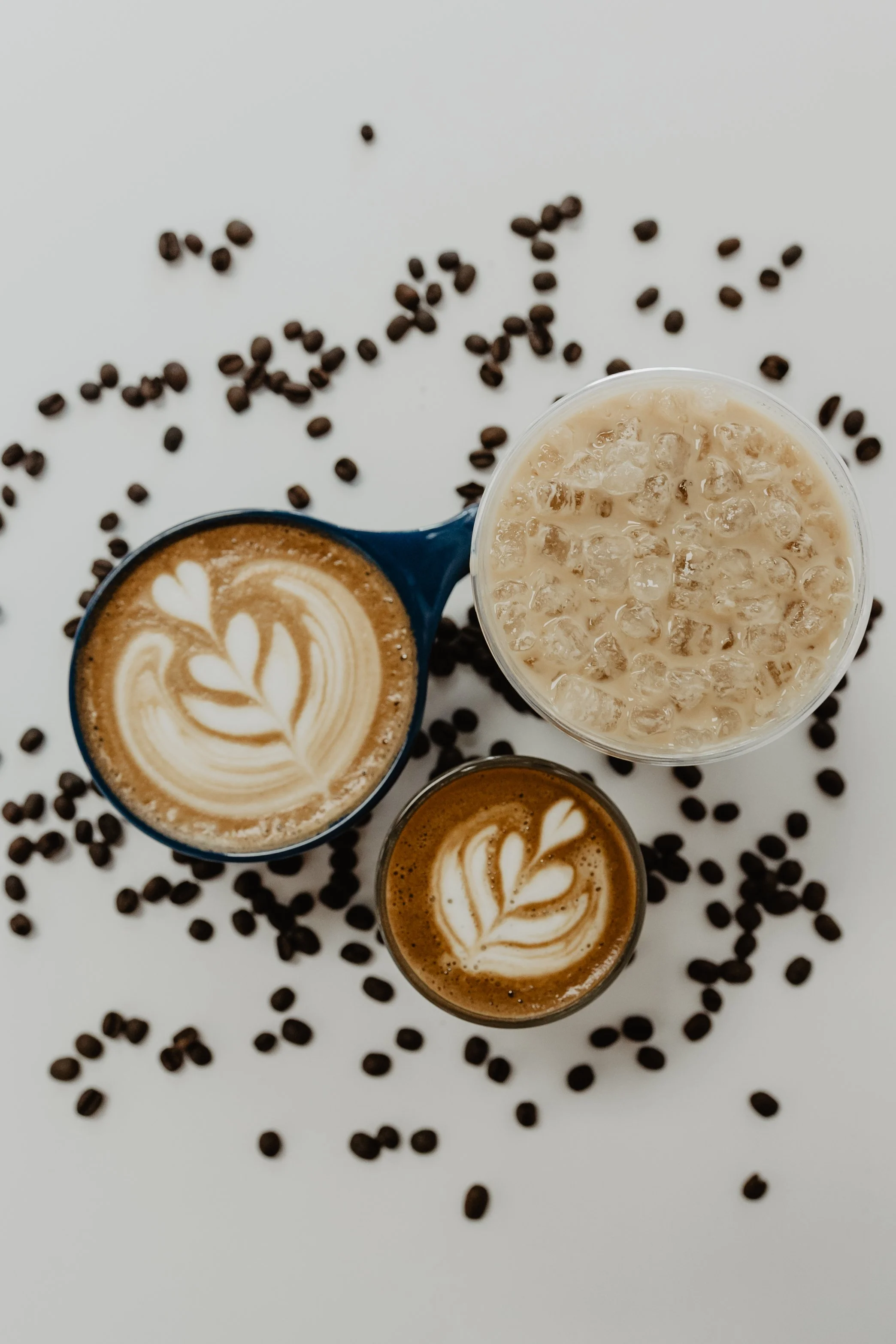 Top view of three coffee cups with latte art on a white surface, surrounded by scattered coffee beans, and a glass of iced coffee.