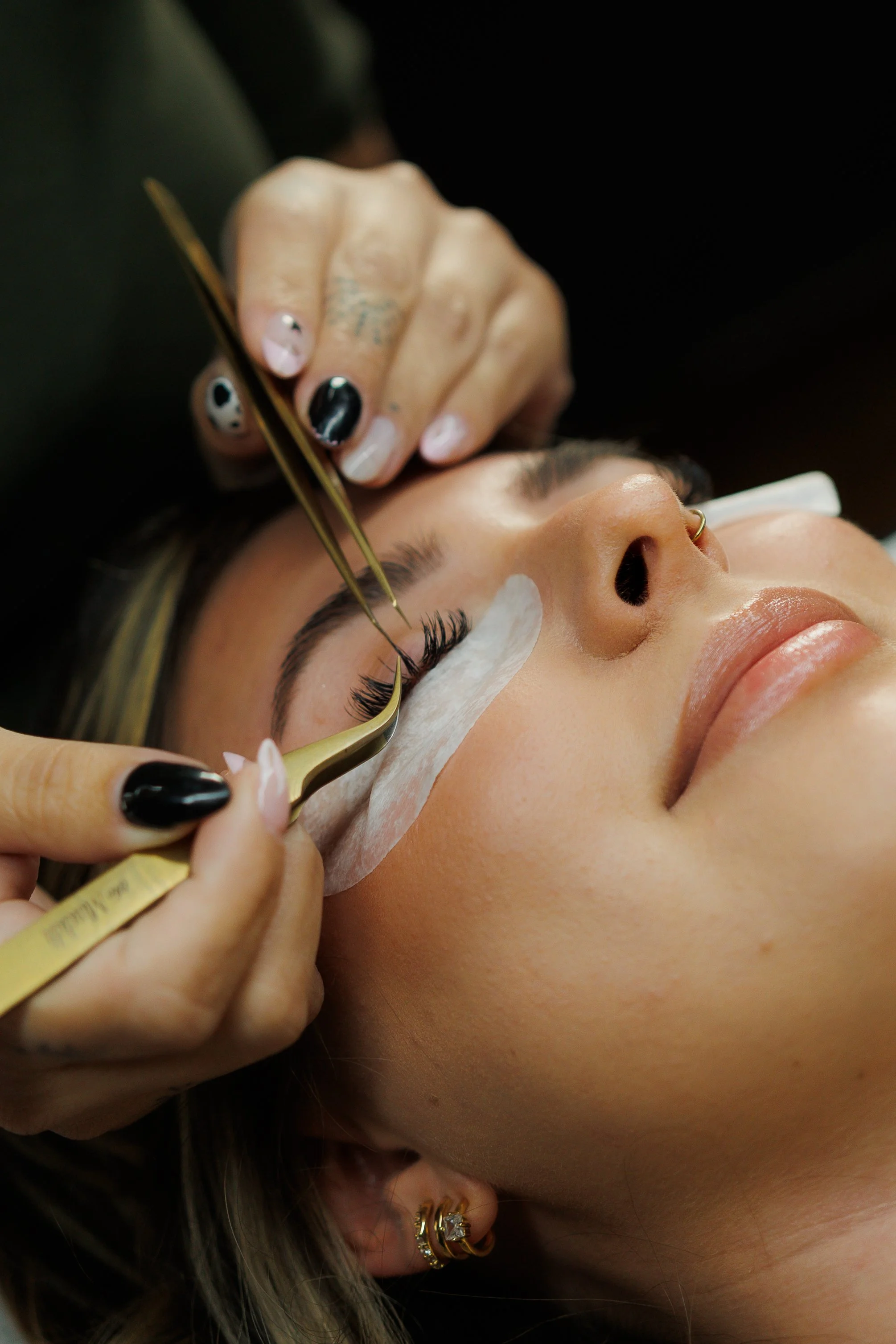A person with a nose ring is receiving eyelash extension treatment. A beauty technician is carefully applying eyelash extensions with a precision tool. The person is lying down with their eyes closed, and the technician's hand is steady and precise.
