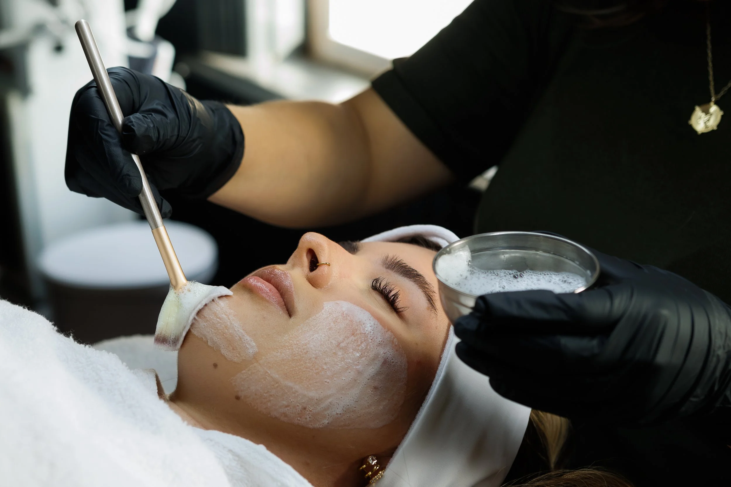 A woman receiving a skincare or facial treatment in a spa or clinic, with a practitioner applying a foam or cleanser to her face.