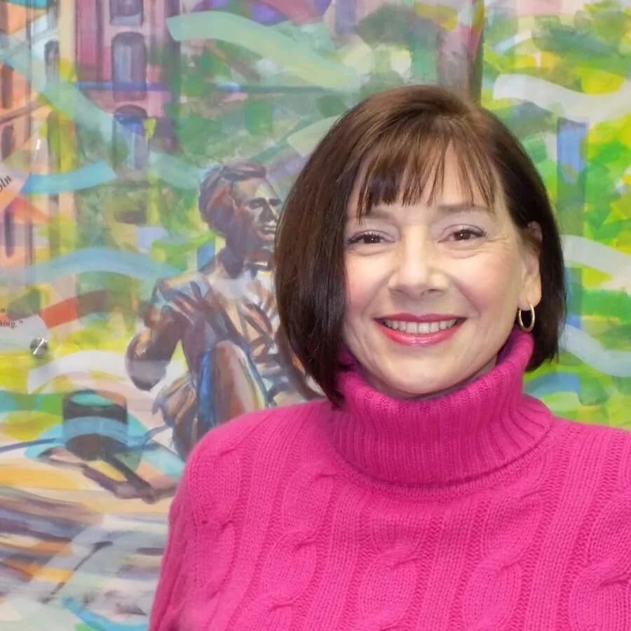 A woman with short brown hair, wearing a bright pink turtleneck sweater and hoop earrings, is smiling at the camera. Behind her is a colorful mural with a portrait of a smiling person and abstract, vibrant cityscape elements.