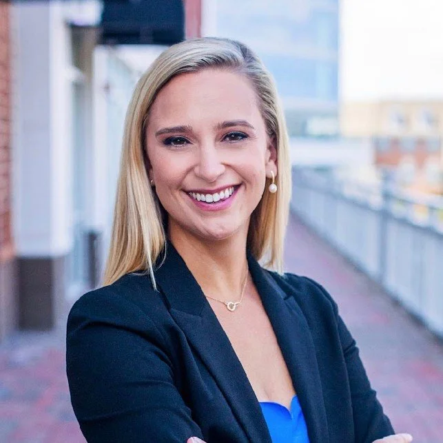 A woman with blonde hair smiling at the camera, wearing a dark blazer and pearl earrings, standing outdoors on a balcony or walkway with city buildings in the background.