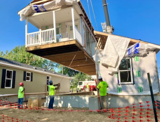 Construction workers inspecting a manufactured home/ modular house being set and lifted by a crane during construction, with blue sky and trees in the background.
