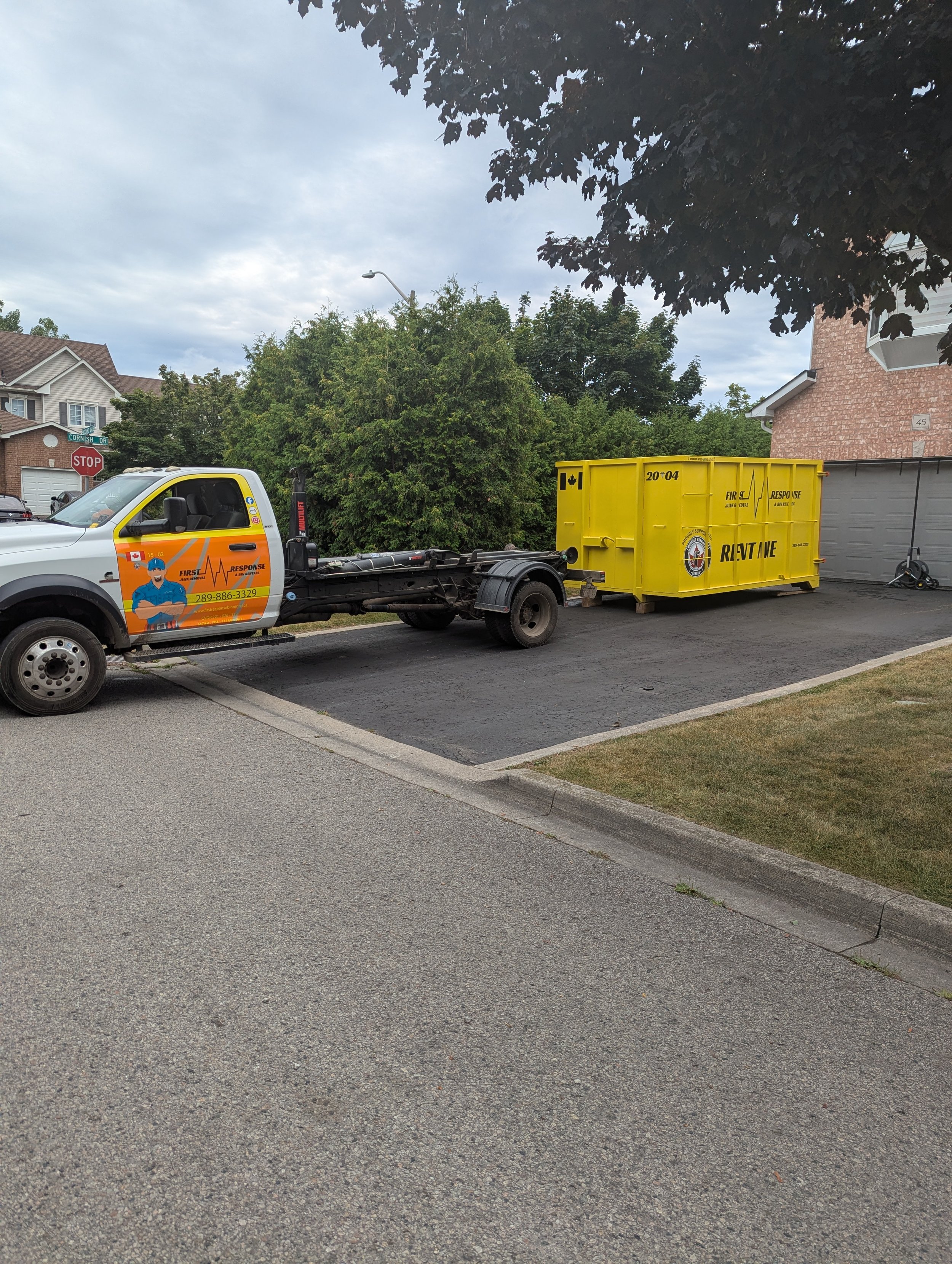 A white response vehicle with a yellow storage container attached in a suburban neighborhood, with houses, trees, and a stop sign visible.
