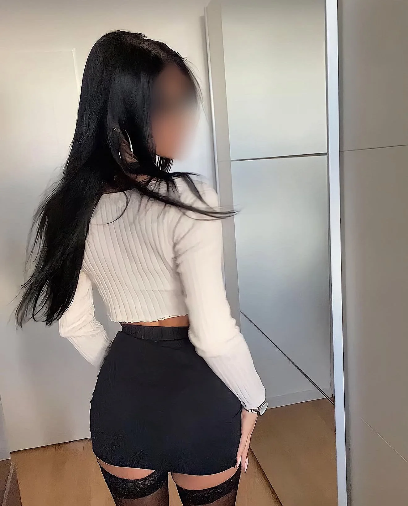 A woman with long black hair standing with her back to the camera, wearing a white long sleeve top, a black skirt, and black thigh-high stockings.