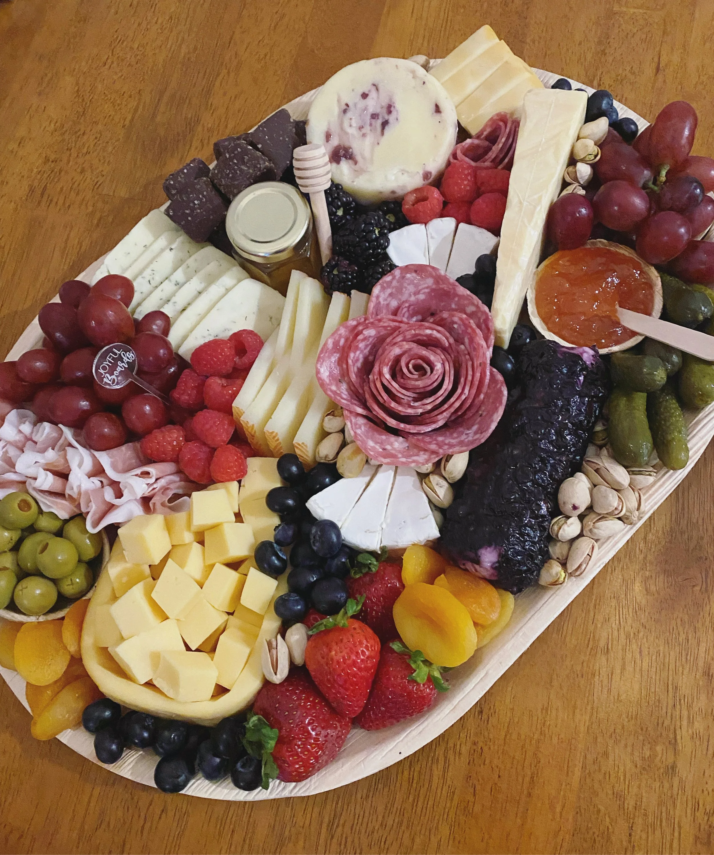 A cheese and fruit platter with various cheeses, grapes, strawberries, raspberries, blackberries, and a honey dipper, arranged on a wooden table.