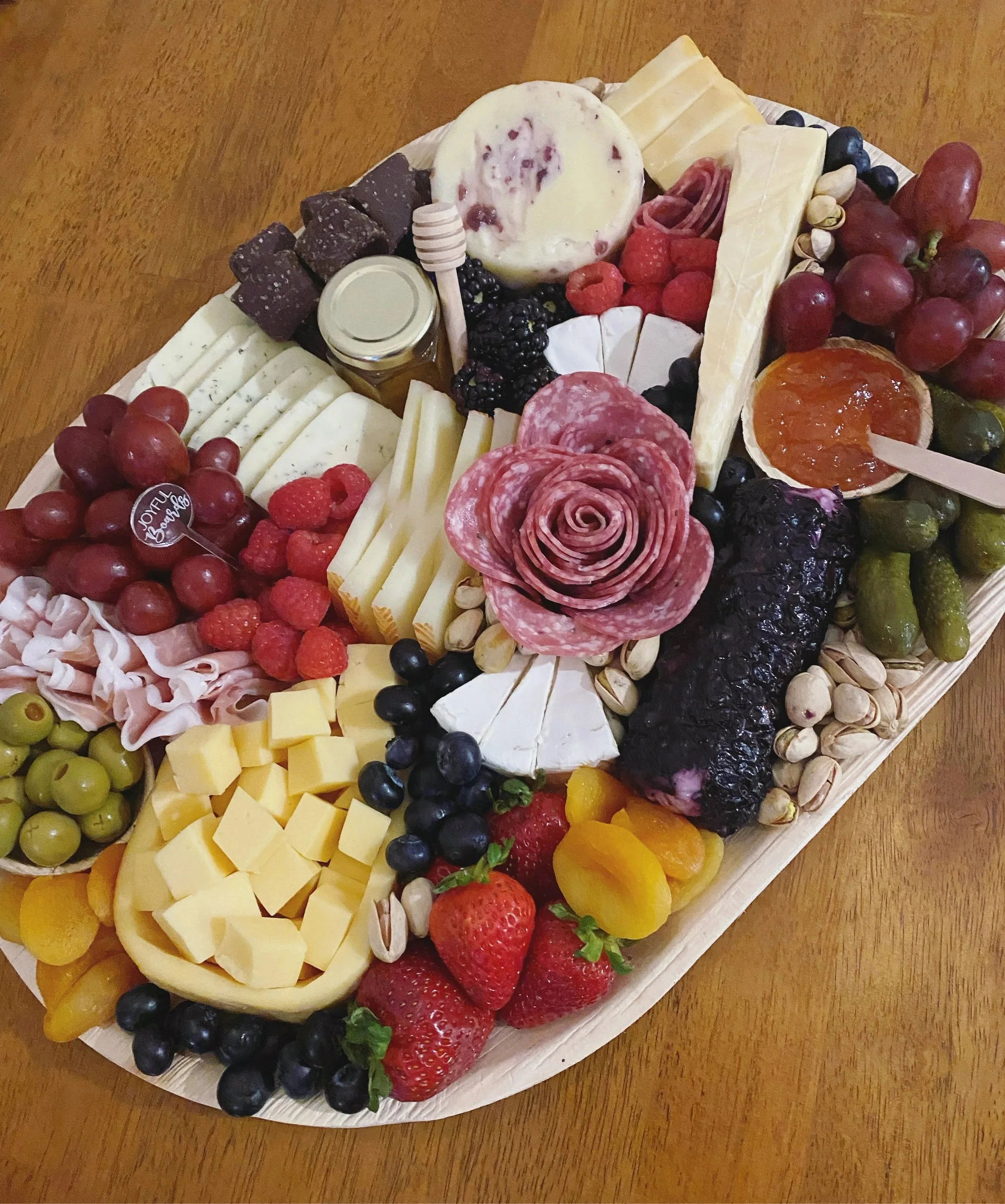 A charcuterie board with assorted cheeses, fresh strawberries, blueberries, blackberries, raspberries, green grapes, red grapes, sliced and rolled meats, a small jar of honey, a round of brie cheese, and a small container of jam, all arranged on a wooden tray.