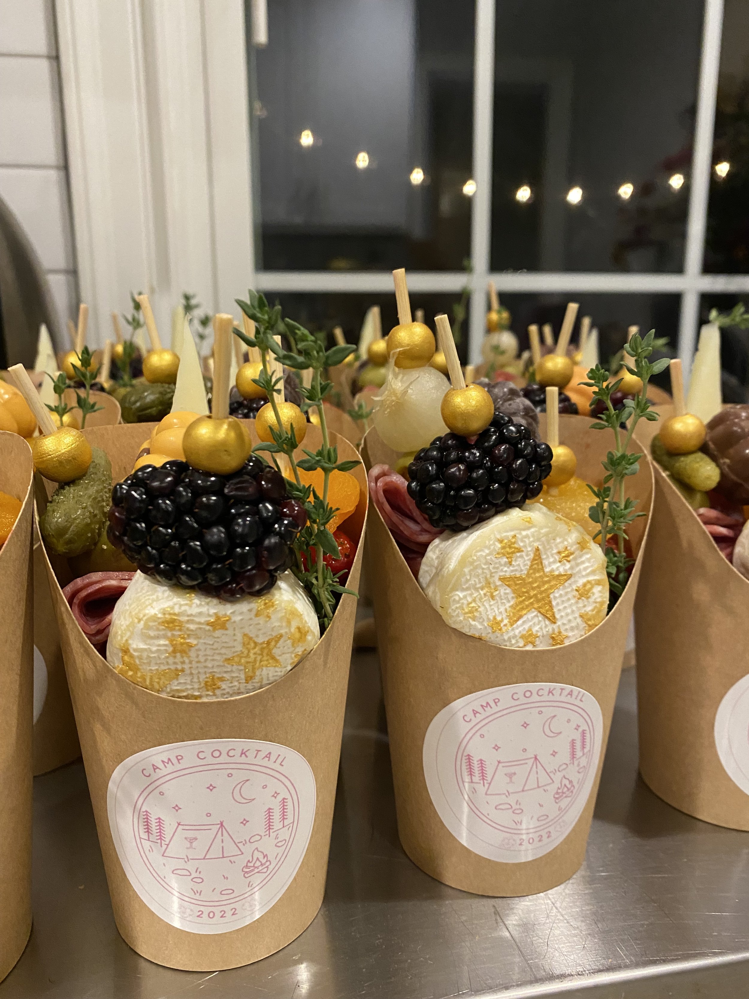 Assorted dessert or snack cups with berries, cheese, and garnishes in paper cones, labeled 'Camp Cocktail' with a camping scene illustration, set on a metal surface.