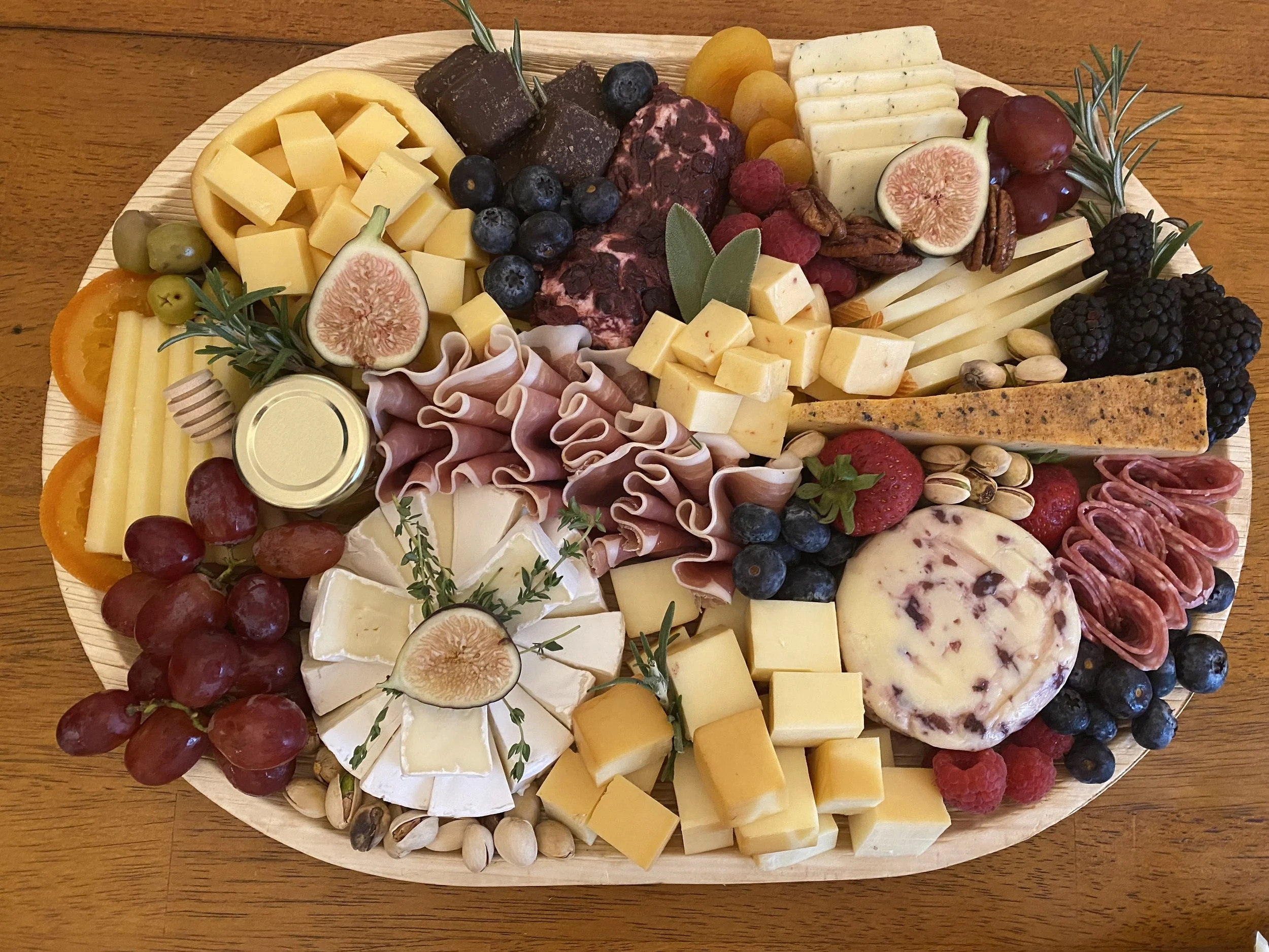A cheese and fruit platter with various cheeses, grapes, figs, berries, nuts, and herbs on a wooden table.