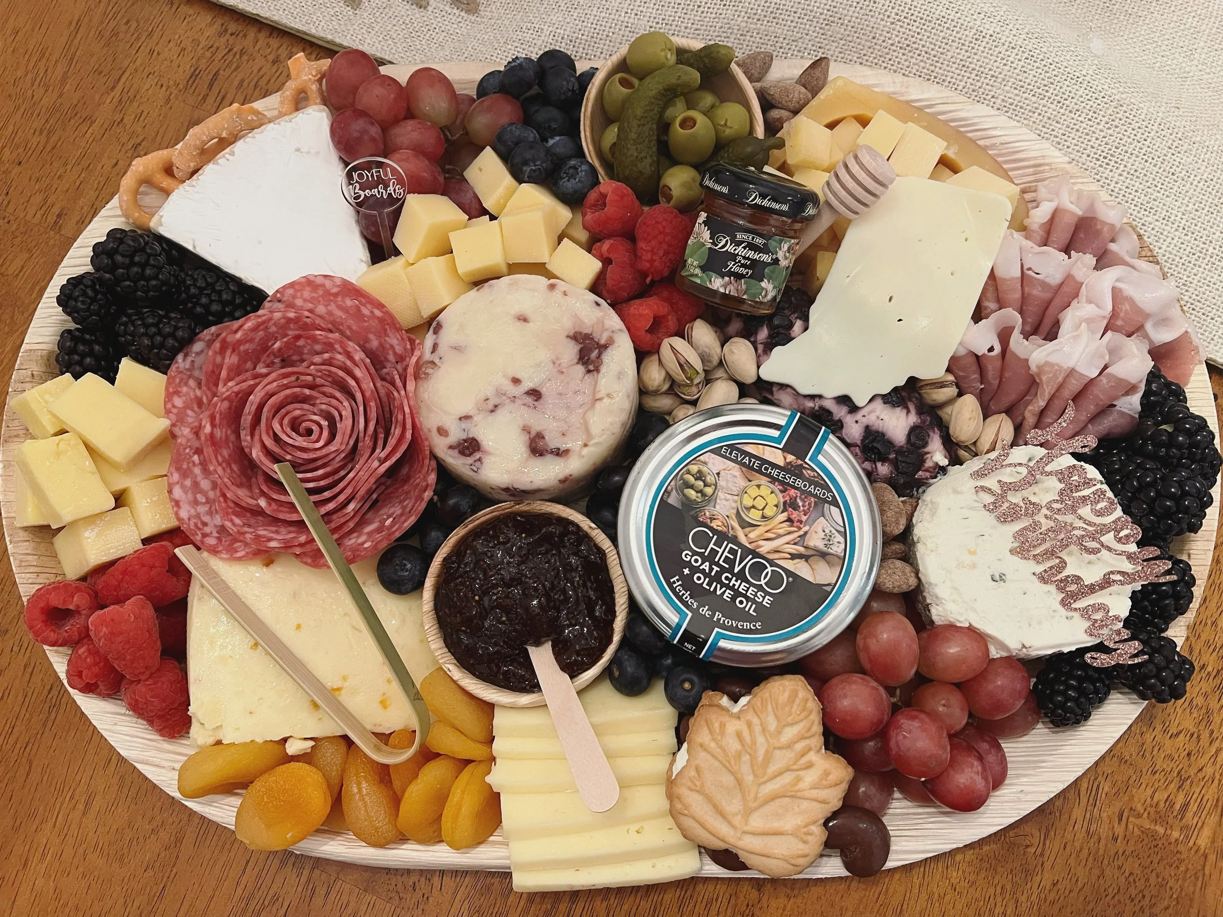 Charcuterie board with assorted cheeses, fresh grapes, blueberries, raspberries, blackberries, olives, nuts, honey, sliced cured meats, and crackers on a wooden table.