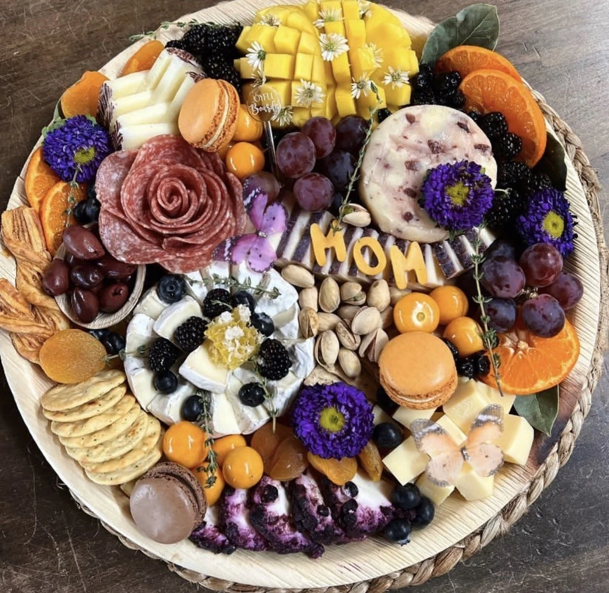 A large round cheese and charcuterie platter with grapes, berries, sliced oranges, mango, blackberries, blueberries, pistachios, crackers, cheese, macarons, and decorative flowers, including a greeting spelling 'MOM' for Mother's Day.