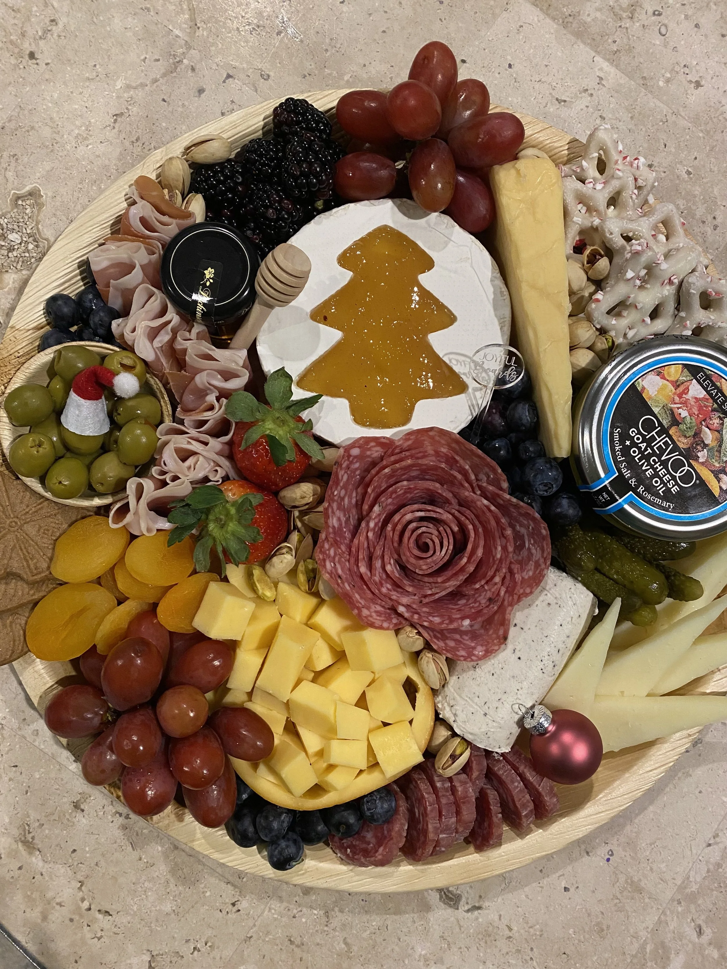 A cheese and charcuterie board with grapes, berries, strawberries, cheese, meats, black olives, nuts, honey, and holiday decorations on a marble surface.