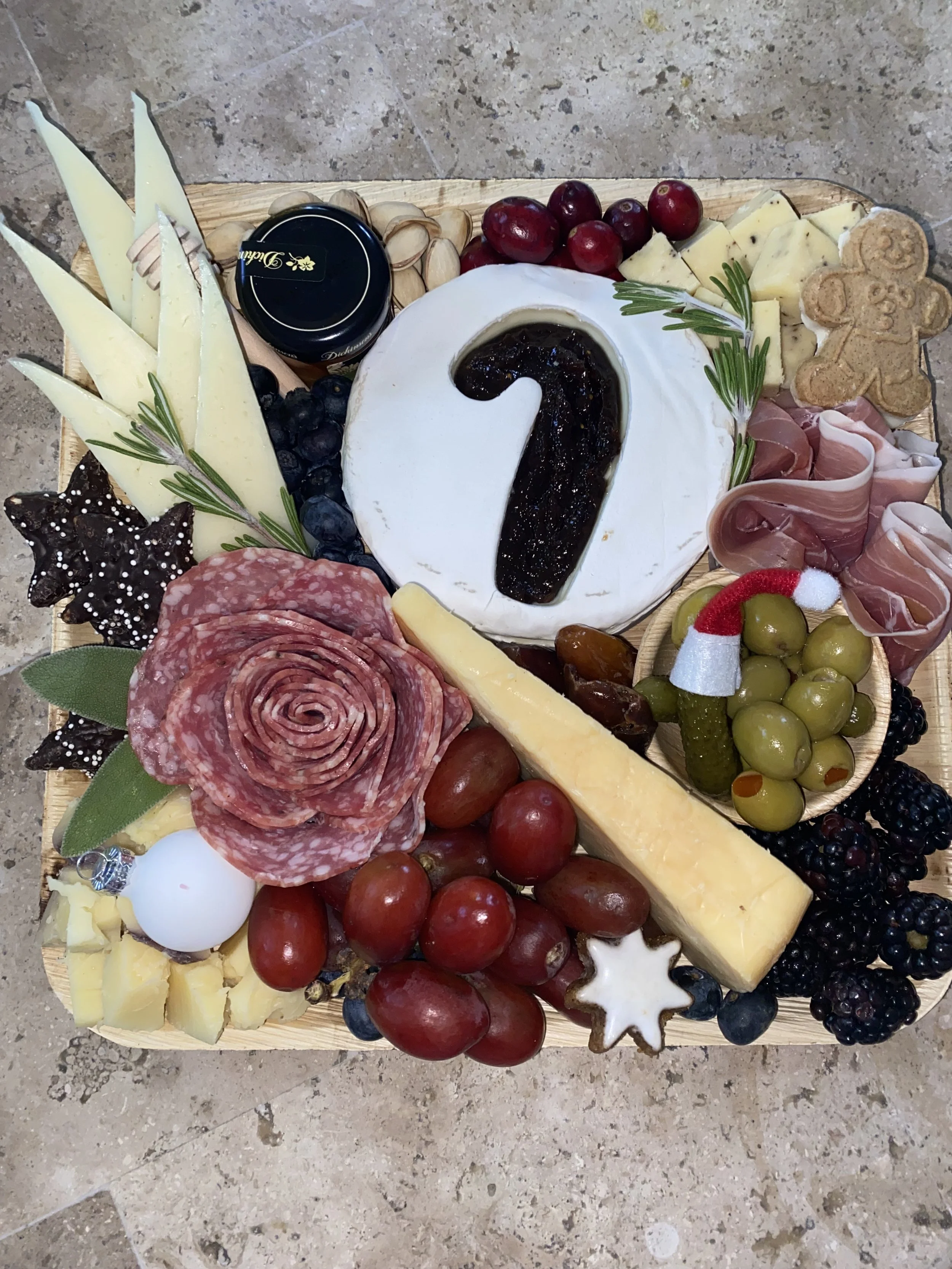A cheese and charcuterie board featuring cheeses, grapes, crackers, meats, cookies, and garnishes.