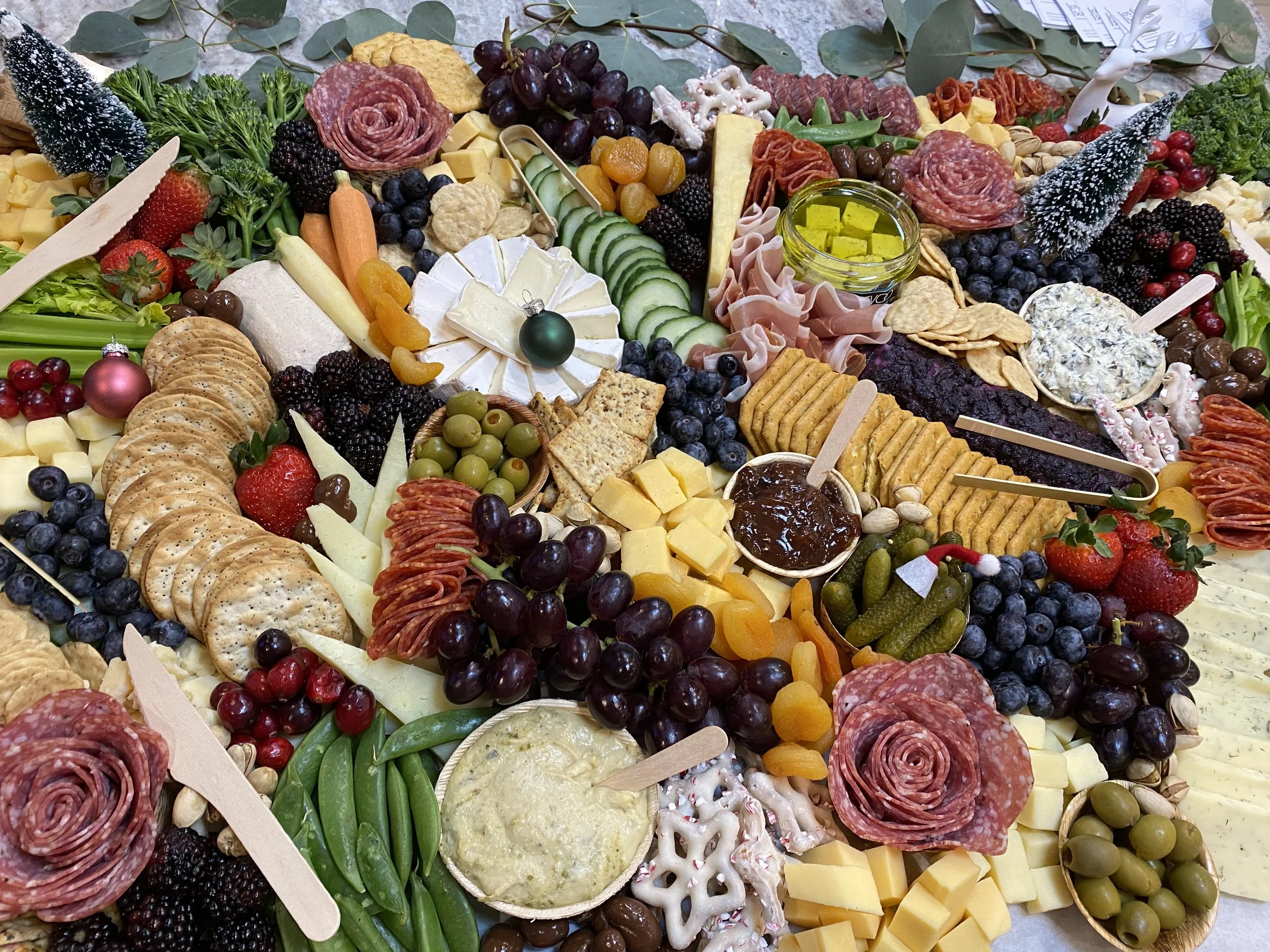 A large charcuterie board with assorted cheeses, meats, crackers, fruits, and garnishes, decorated with small Christmas trees and holiday-themed items.