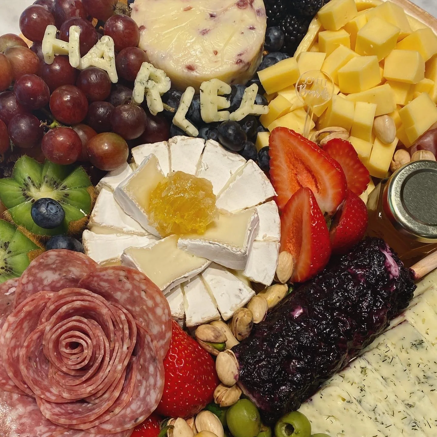 Assorted cheese, grapes, strawberries, kiwi, blackberries, blueberries, sliced strawberries, sliced cheese, honey jar, pistachios, cured meats, jam, and herbs.
