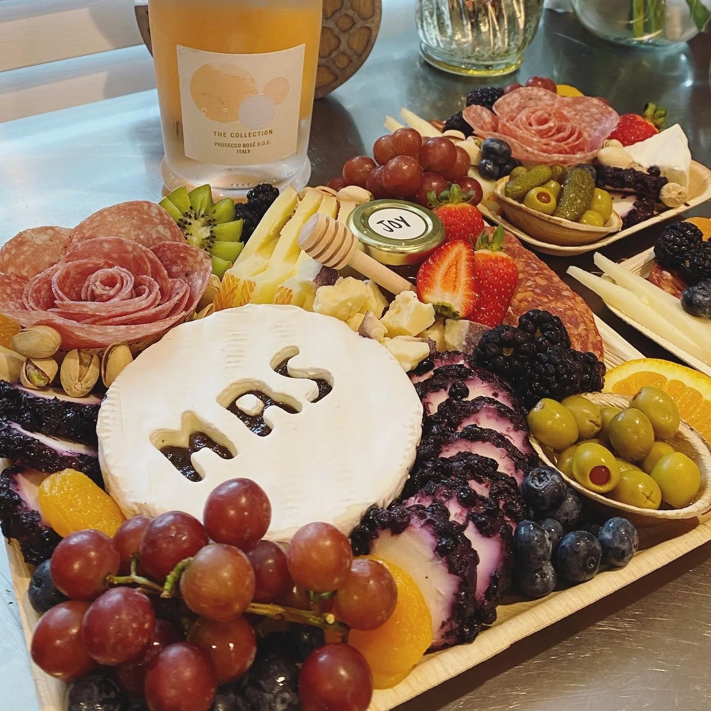 A cheese and charcuterie board with grapes, strawberries, blueberries, blackberries, sliced oranges, and a wheel of Brie cheese with the word 'LOVE' cut out. There is also sliced cured meats, nuts, and small bowls of pickles and jam. A bottle of Pros