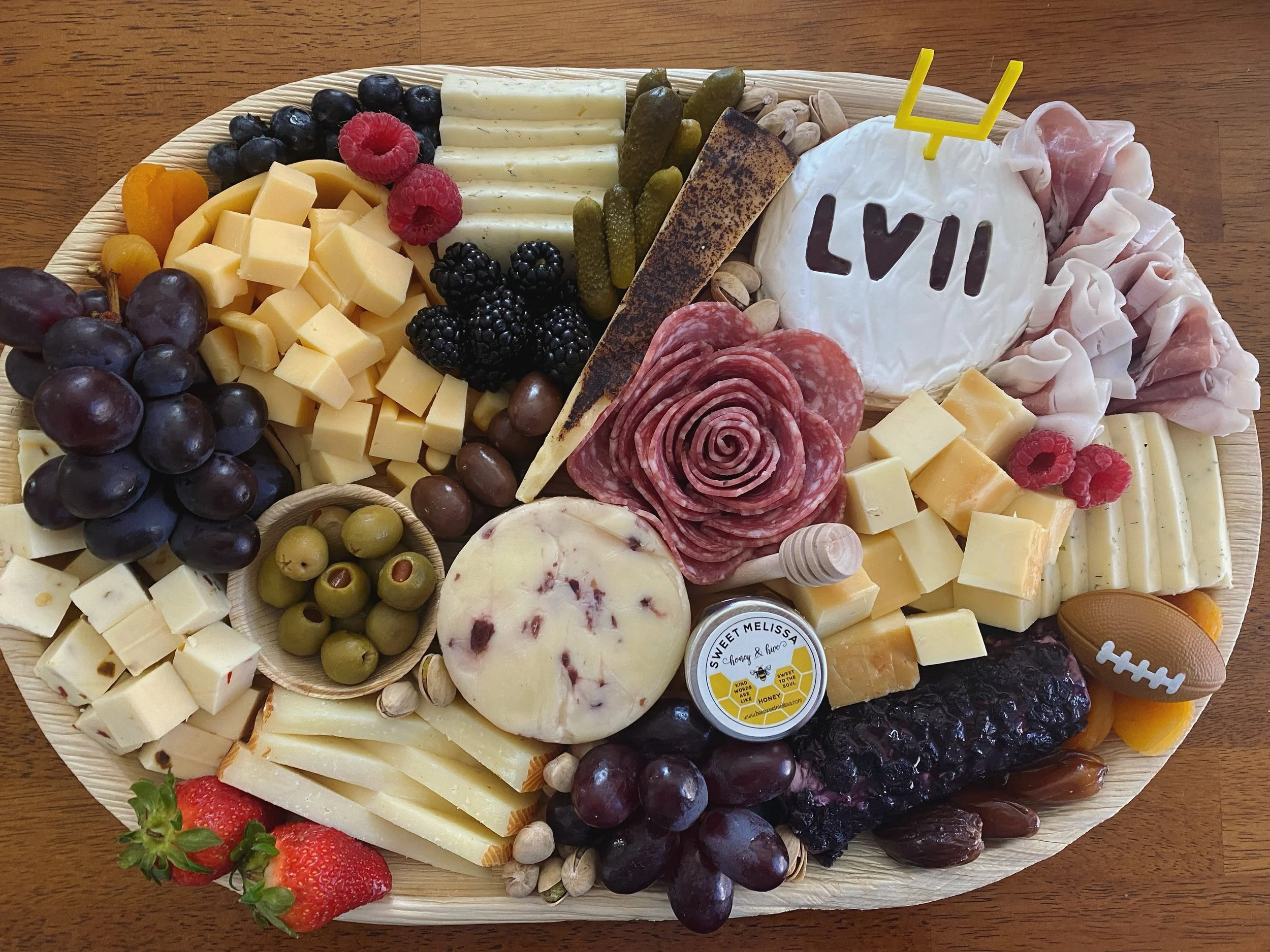 A cheese and fruit platter with grapes, raspberries, strawberries, cheese cubes, sliced cheese, pickles, nuts, and meats on a round wooden table.