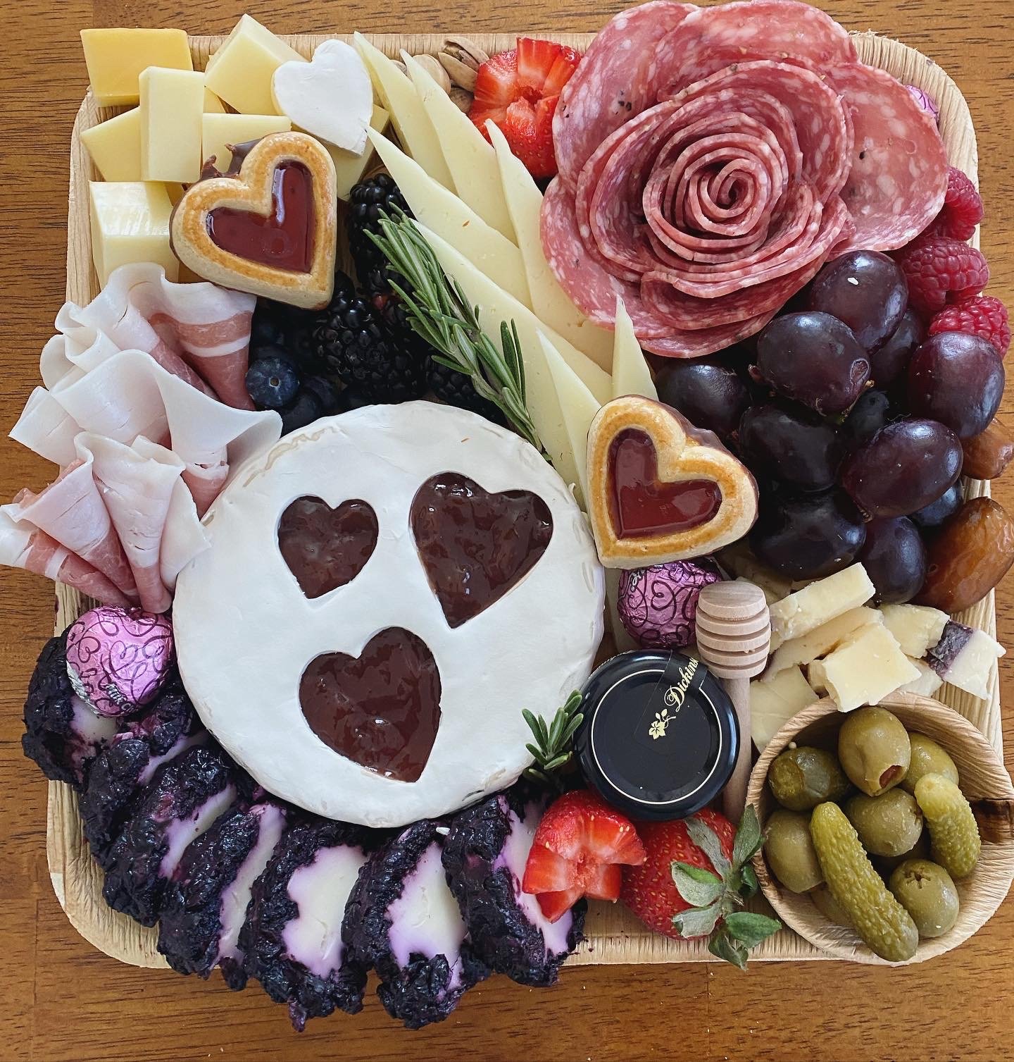 A wooden serving platter with assorted cheeses, meats, fruits, chocolate, and crackers, including strawberries, blackberries, grapes, grapes, pineapple, and strawberries, with heart-shaped chocolates, a cheese wheel with heart cutouts, berry-topped c