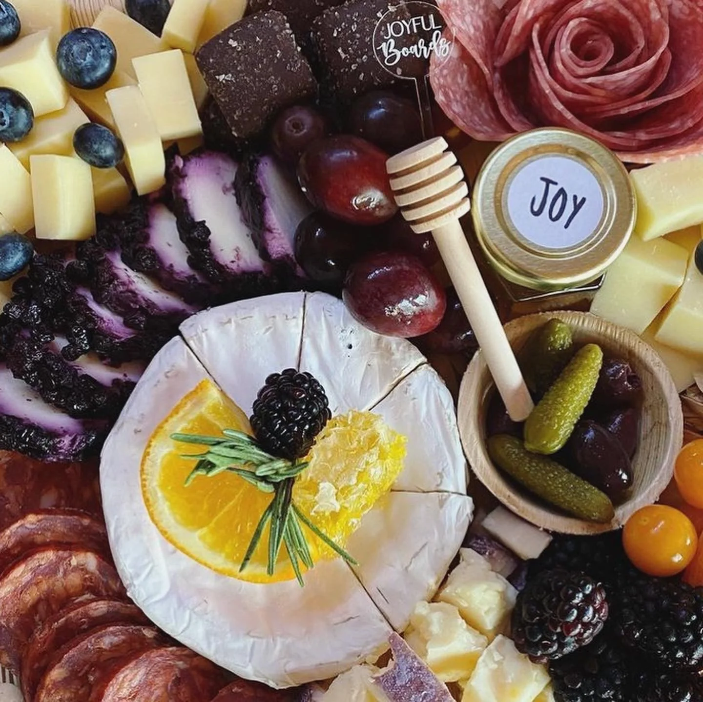 A close-up of a cheese platter featuring brie cheese topped with orange slices, blackberries, and rosemary, alongside grapes, honey, assorted cheeses, crackers, pickles, and slices of meats and fruits.