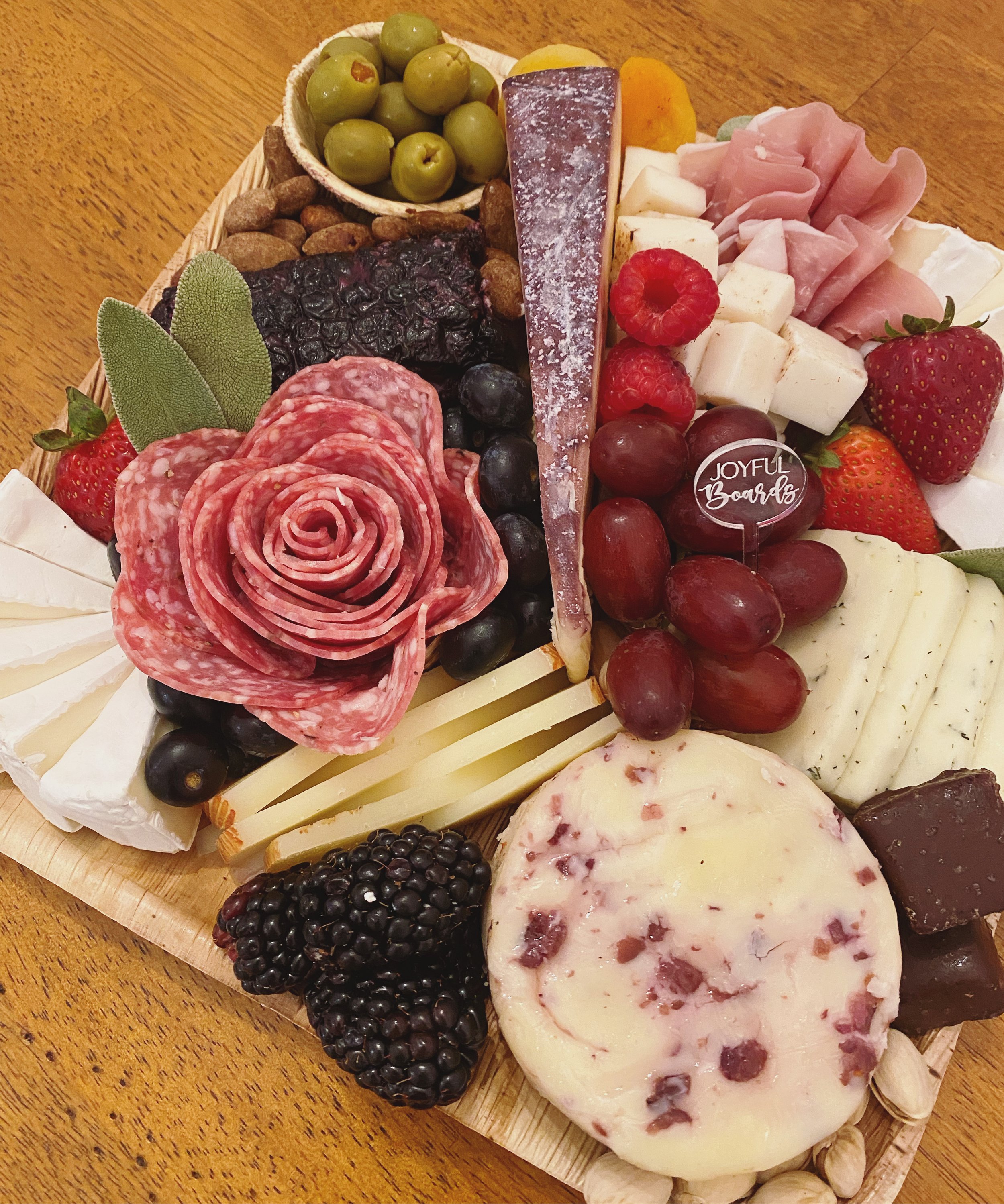 A cheese board with grapes, nuts, dried fruit, cured meats, berries, various cheeses, and herbs arranged on a wooden surface.