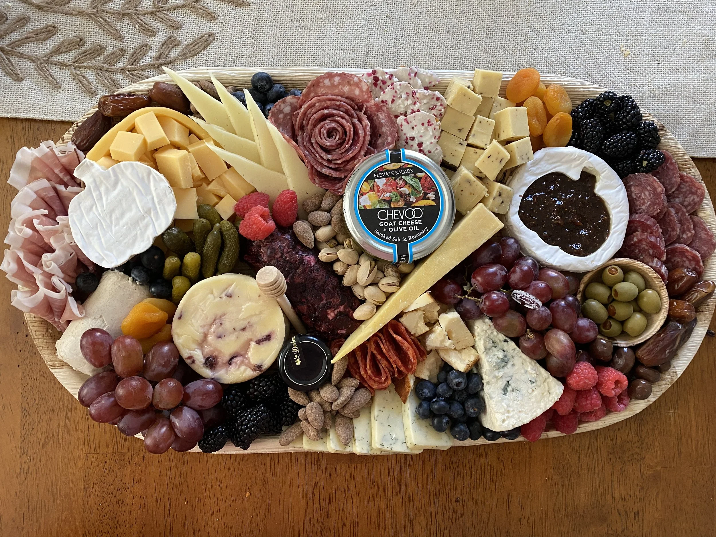 A cheese and charcuterie platter with various cheeses, meats, grapes, berries, crackers, and a jar of goat cheese with herbs, arranged on a wooden tray.