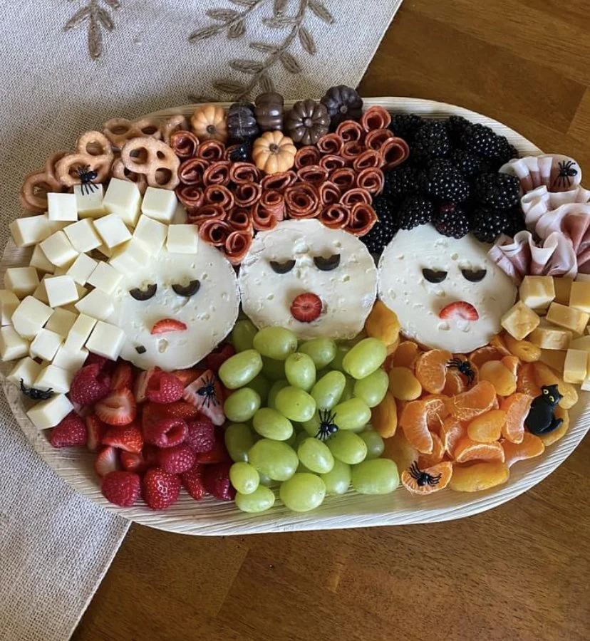 Fruit and cheese platter arranged in the shape of a rainbow, with three smiling faces, strawberries, grapes, blackberries, orange slices, and various cheeses.