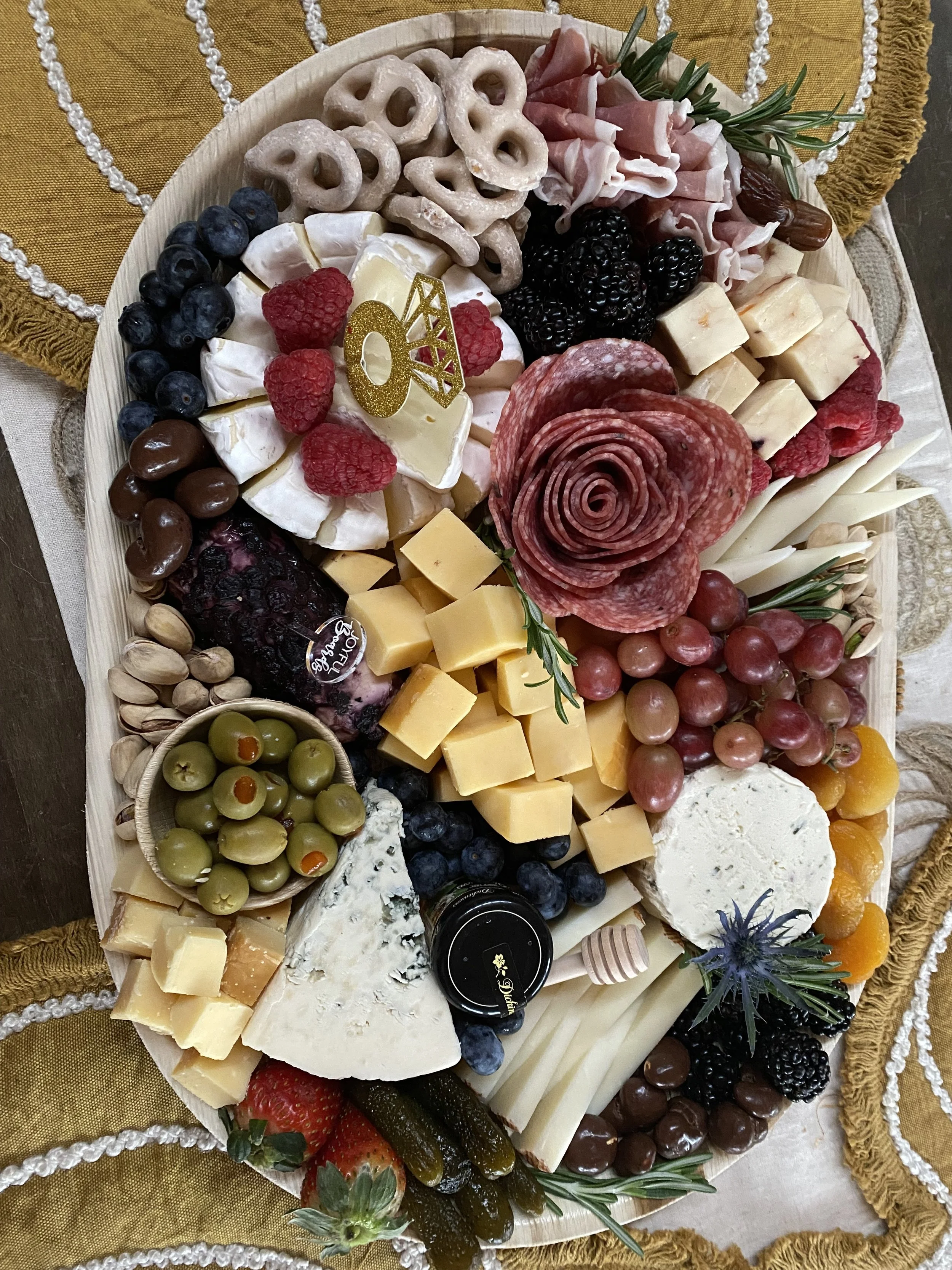 An elaborate cheese and charcuterie board with grapes, berries, nuts, cheese, cured meats, and crackers, garnished with herbs and flowers, on a decorative table setting.