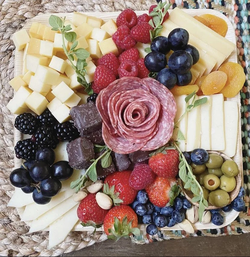 A cheese and fruit platter with various cheeses, fresh berries, sliced peaches, green olives, dried apricots, salami slices, and garnished with sprigs of fresh herbs.