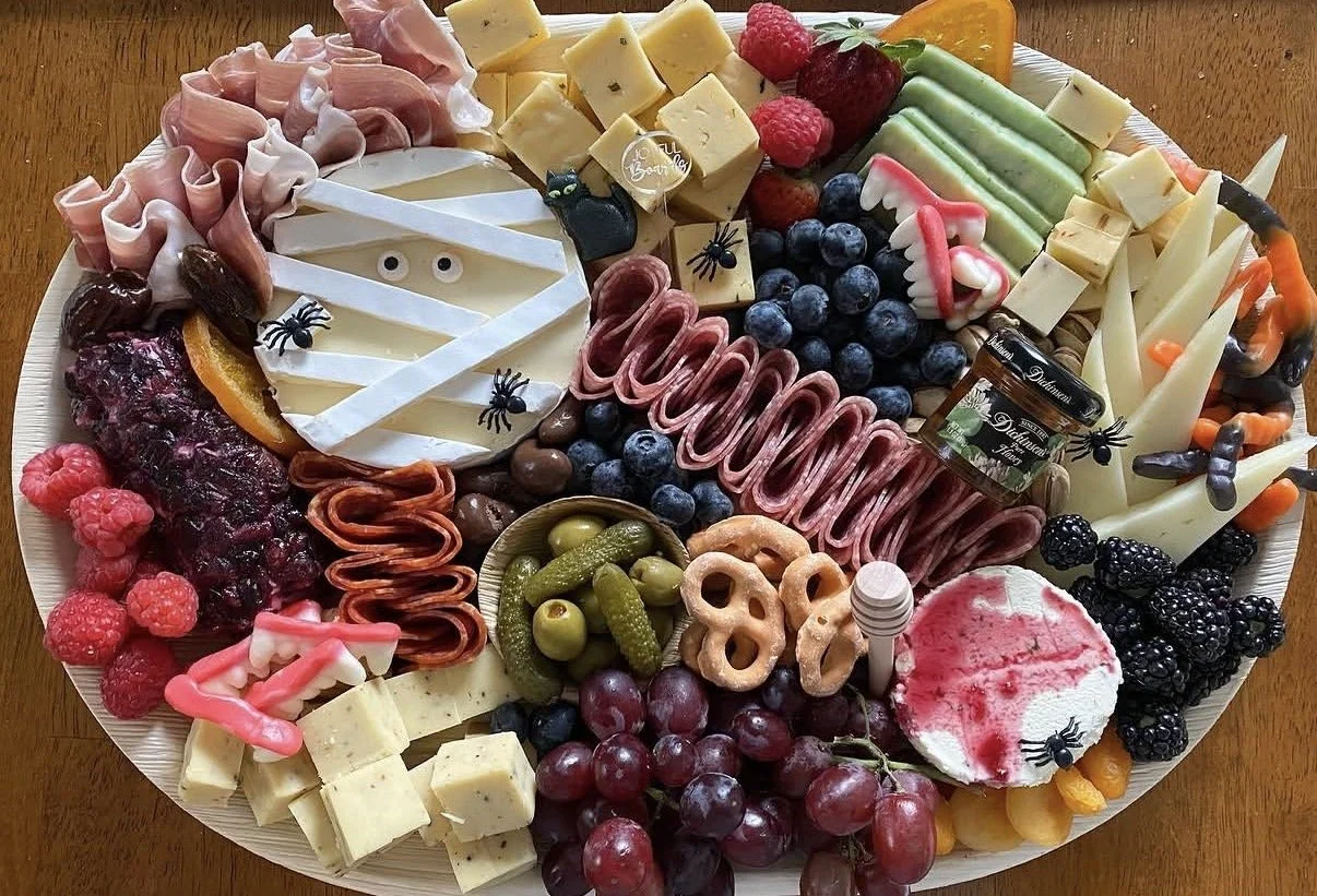 A Halloween-themed cheese and fruit platter with assorted cheeses, grapes, berries, sliced fruits, and decorative Halloween edible embellishments like plastic spiders and cobwebs.
